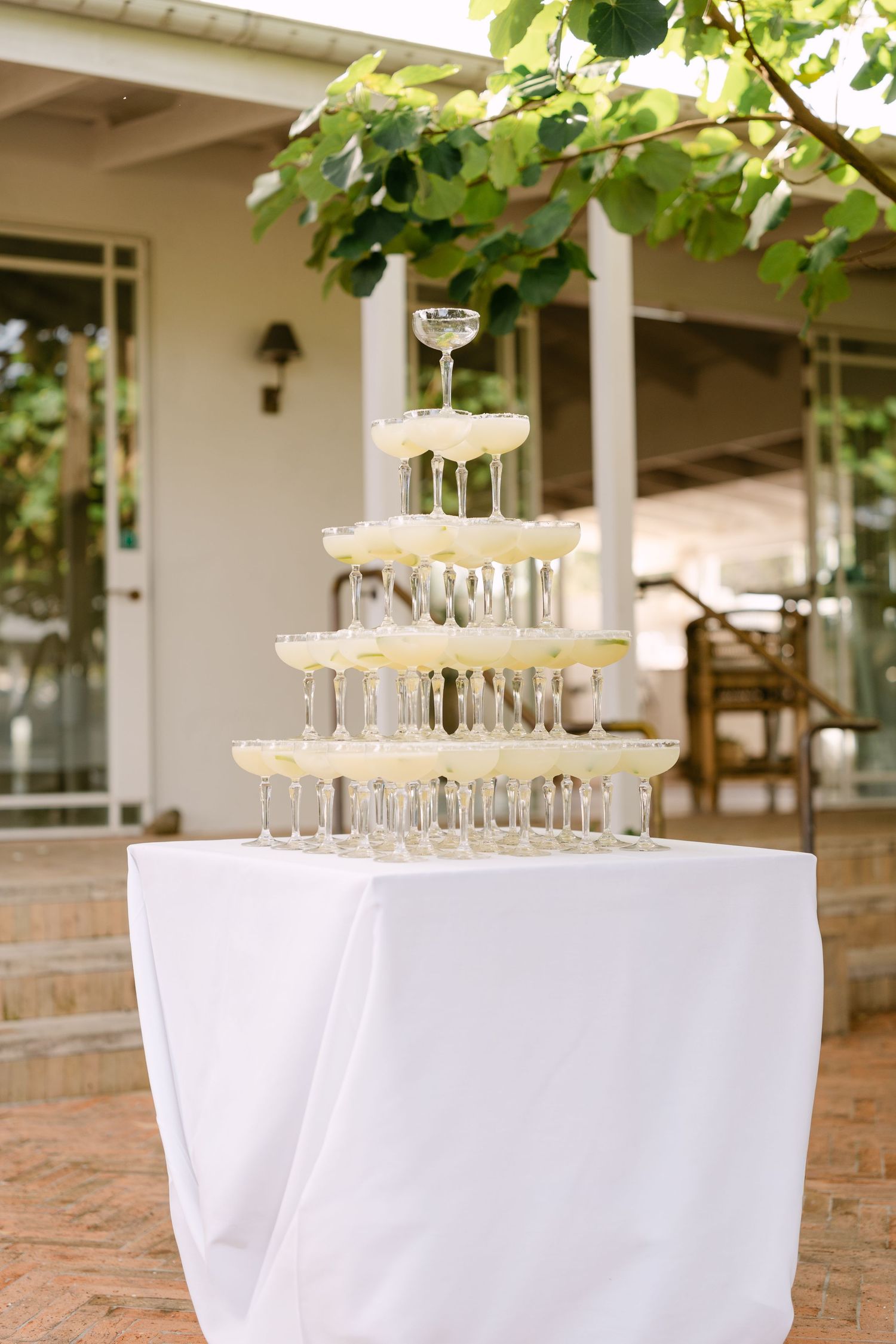 Champagne coupe tower stacked on a white-draped table on a sunlit patio beneath green leaves, ideal for wedding receptions.