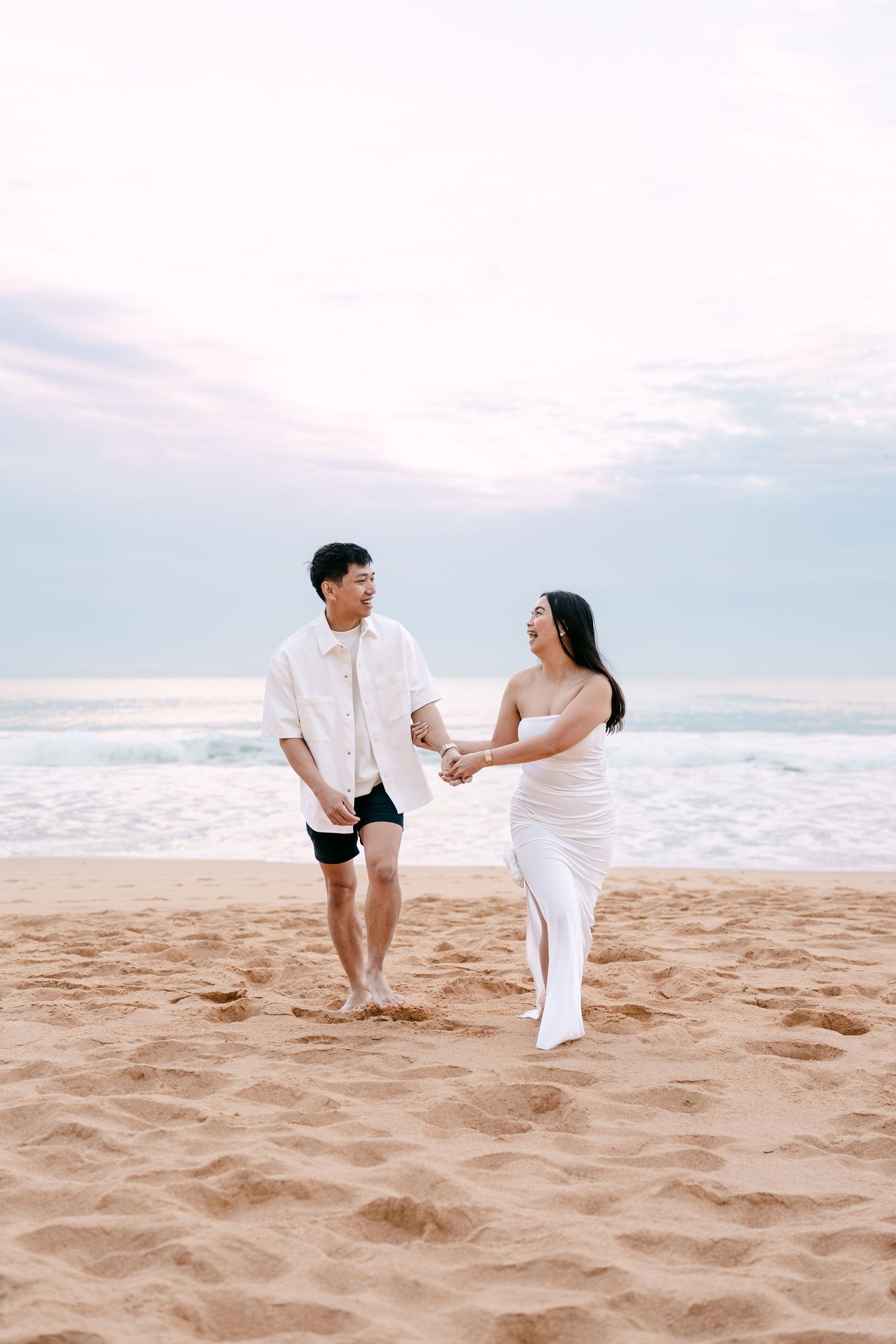 Asian couple holding hands and smiling while walking on sandy beach at dusk.