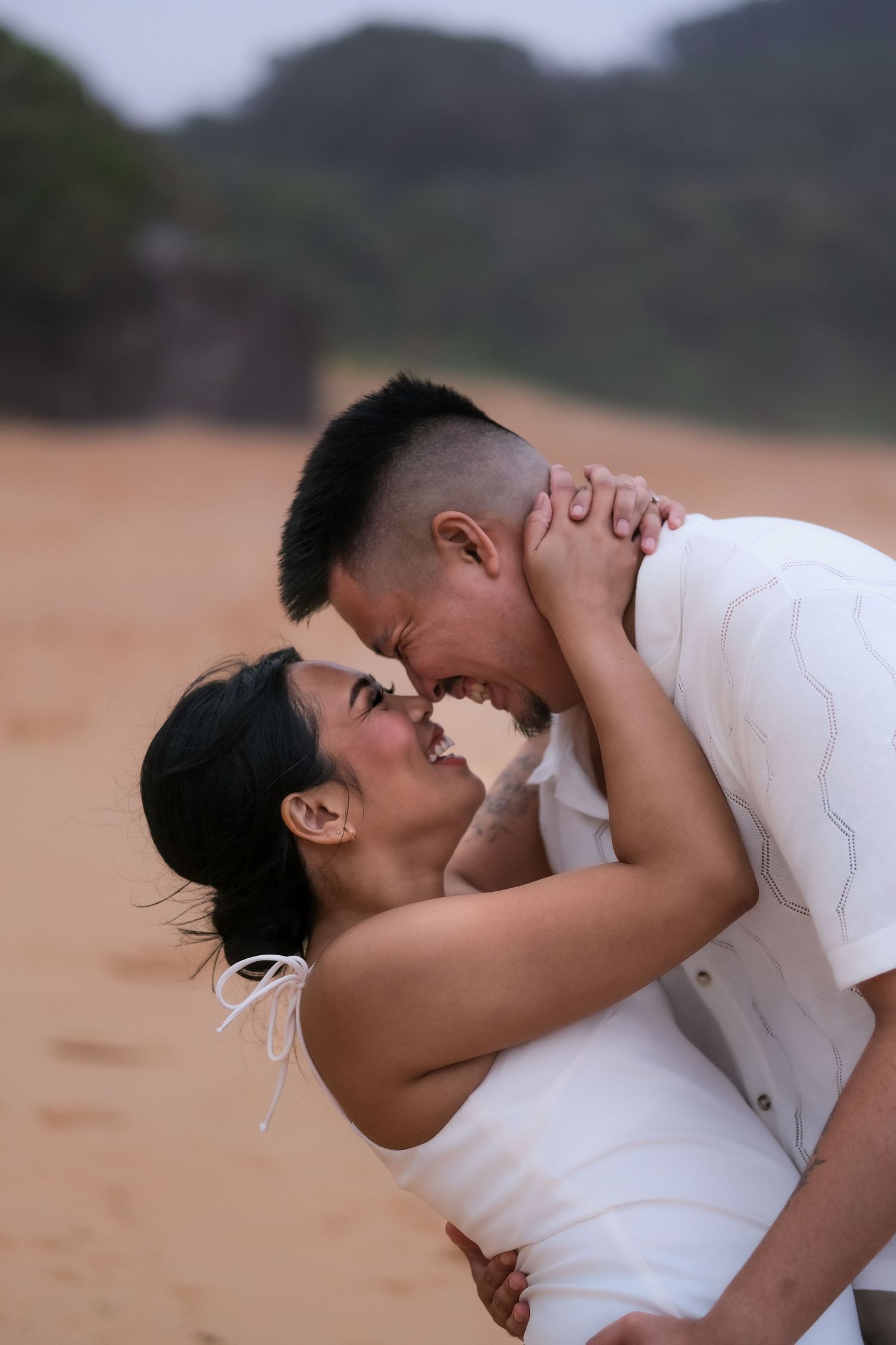 Joyful couple laughing and embracing closely on a sandy beach with misty hills behind.