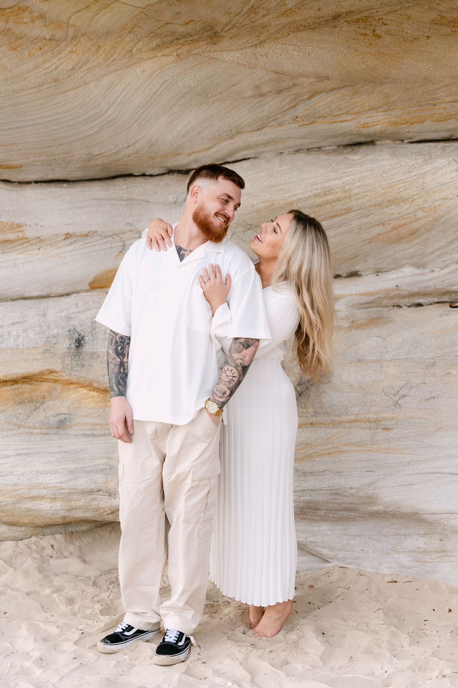 Tattooed couple in white outfits embracing against a sandstone rock wall on a beach.