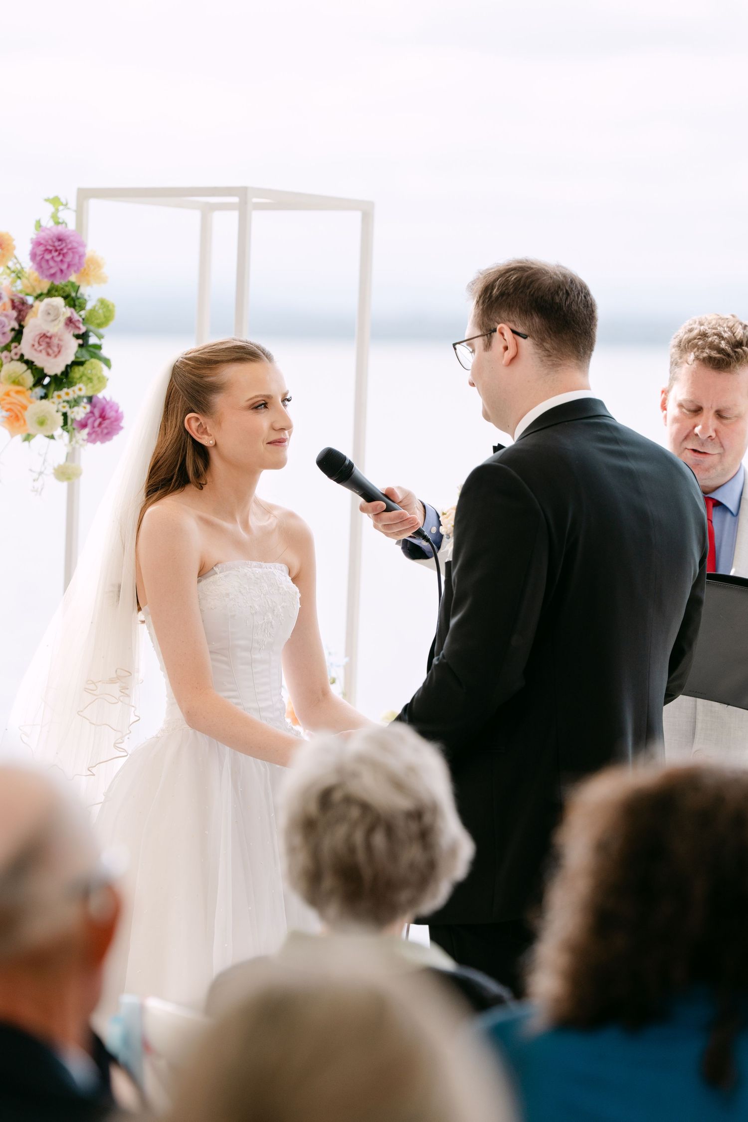 Bride in a strapless white gown and veil faces the groom as an officiant holds a microphone during an outdoor lakeside wedding ceremony.