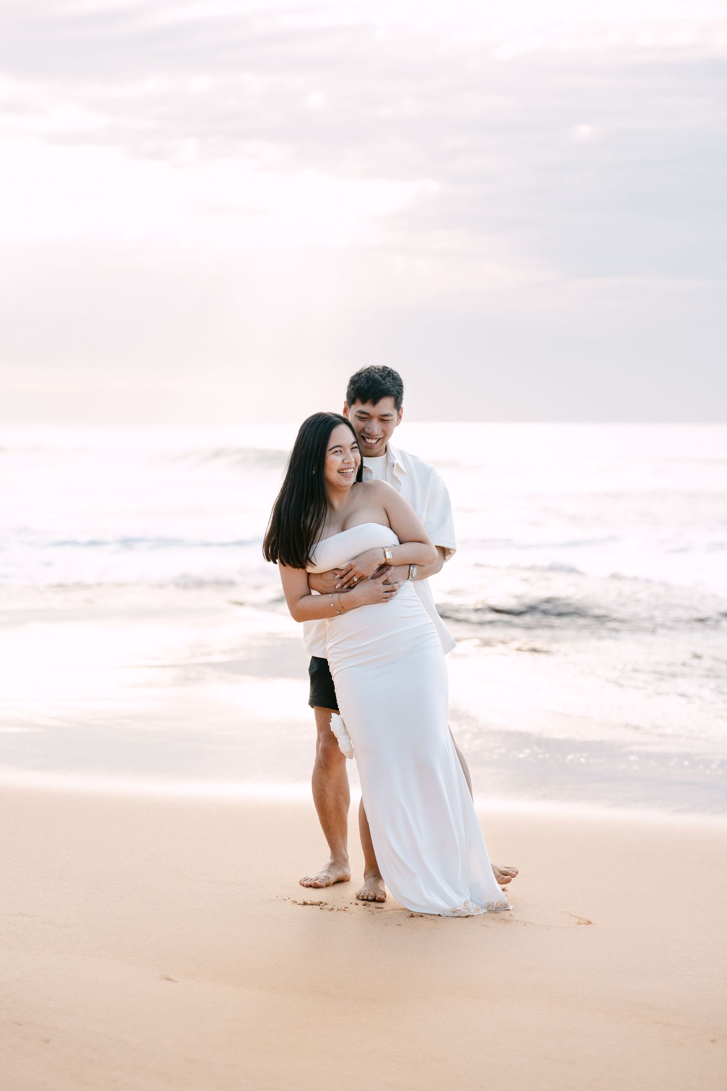 Romantic couple hugging on beach at sunset, woman in white maxi dress, man in white shirt, ocean waves behind them.