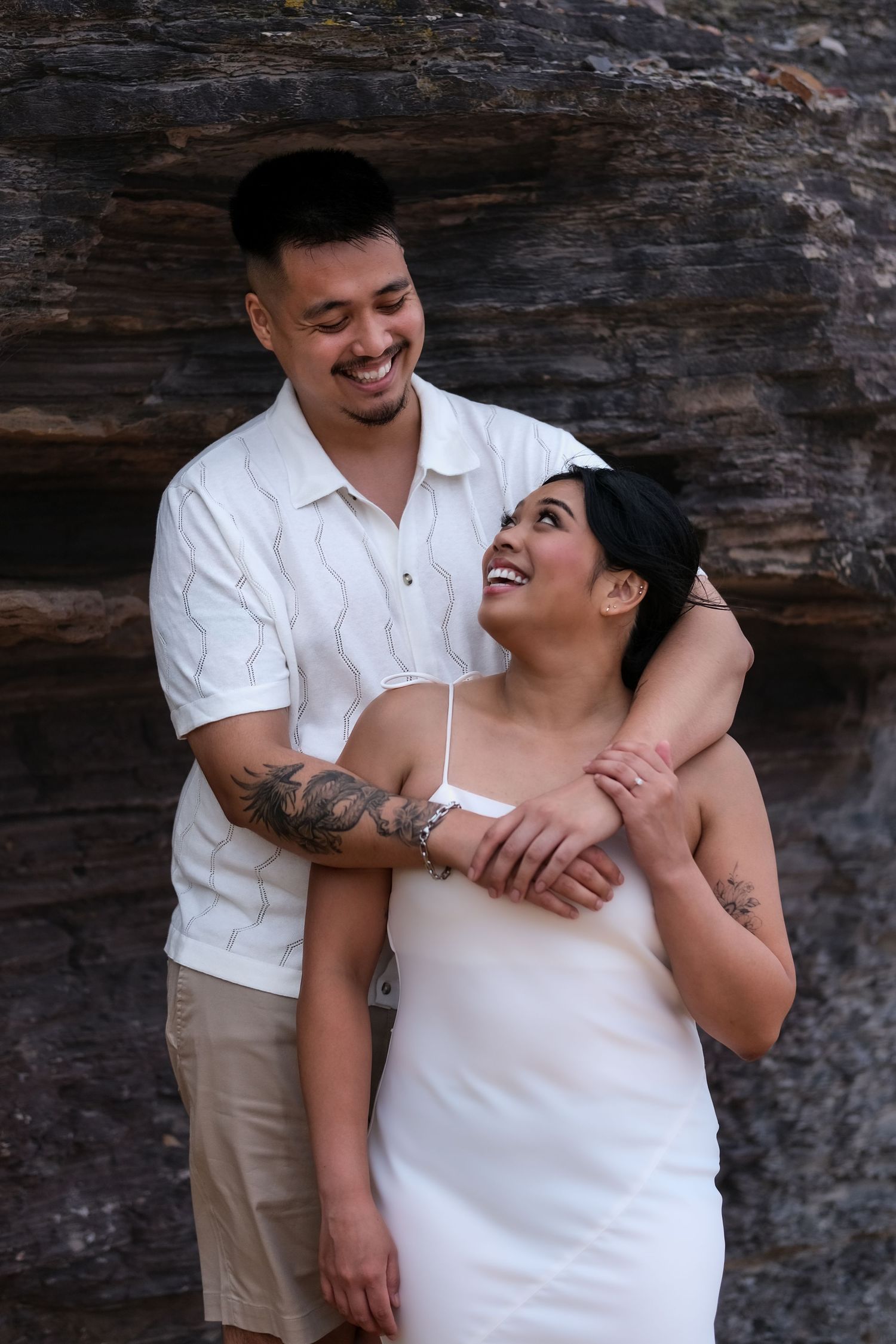 Smiling couple embracing against a rock wall, woman in white dress with tattoos visible.