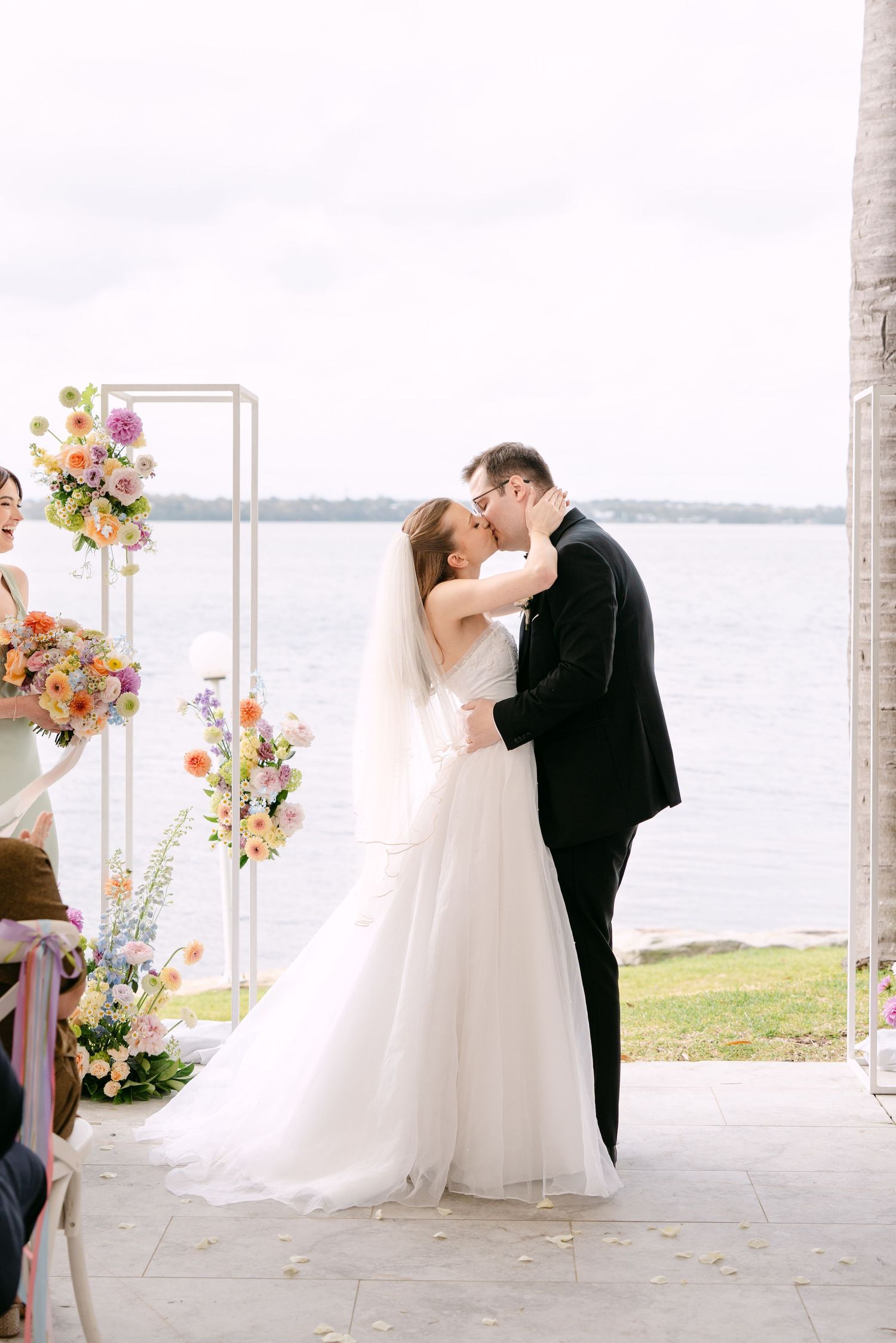 Bride and groom share a first kiss at a waterfront wedding ceremony beside pastel floral arrangements and a white arch.