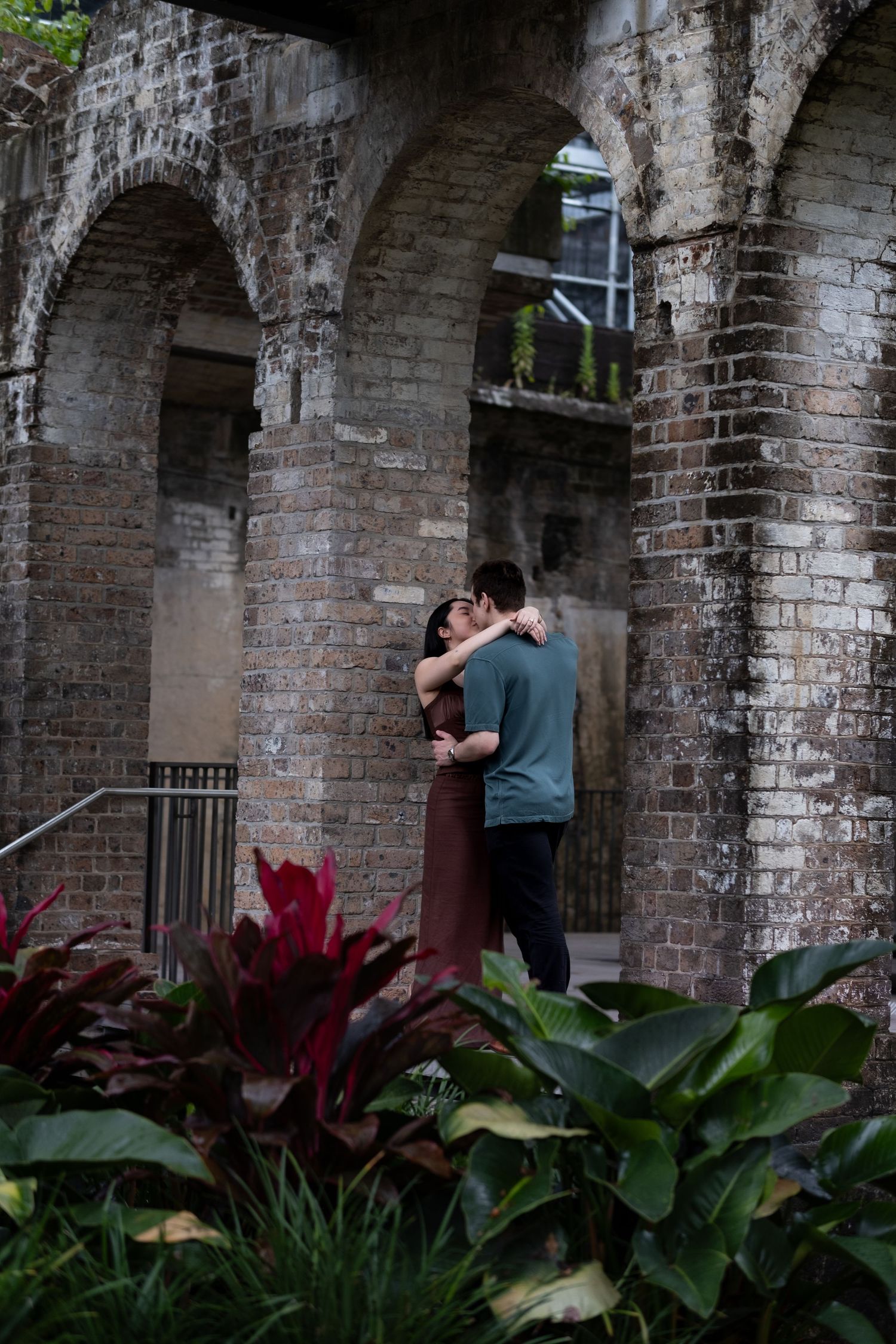 Couple embracing romantically among rustic brick arches and lush tropical plants in an outdoor garden setting.