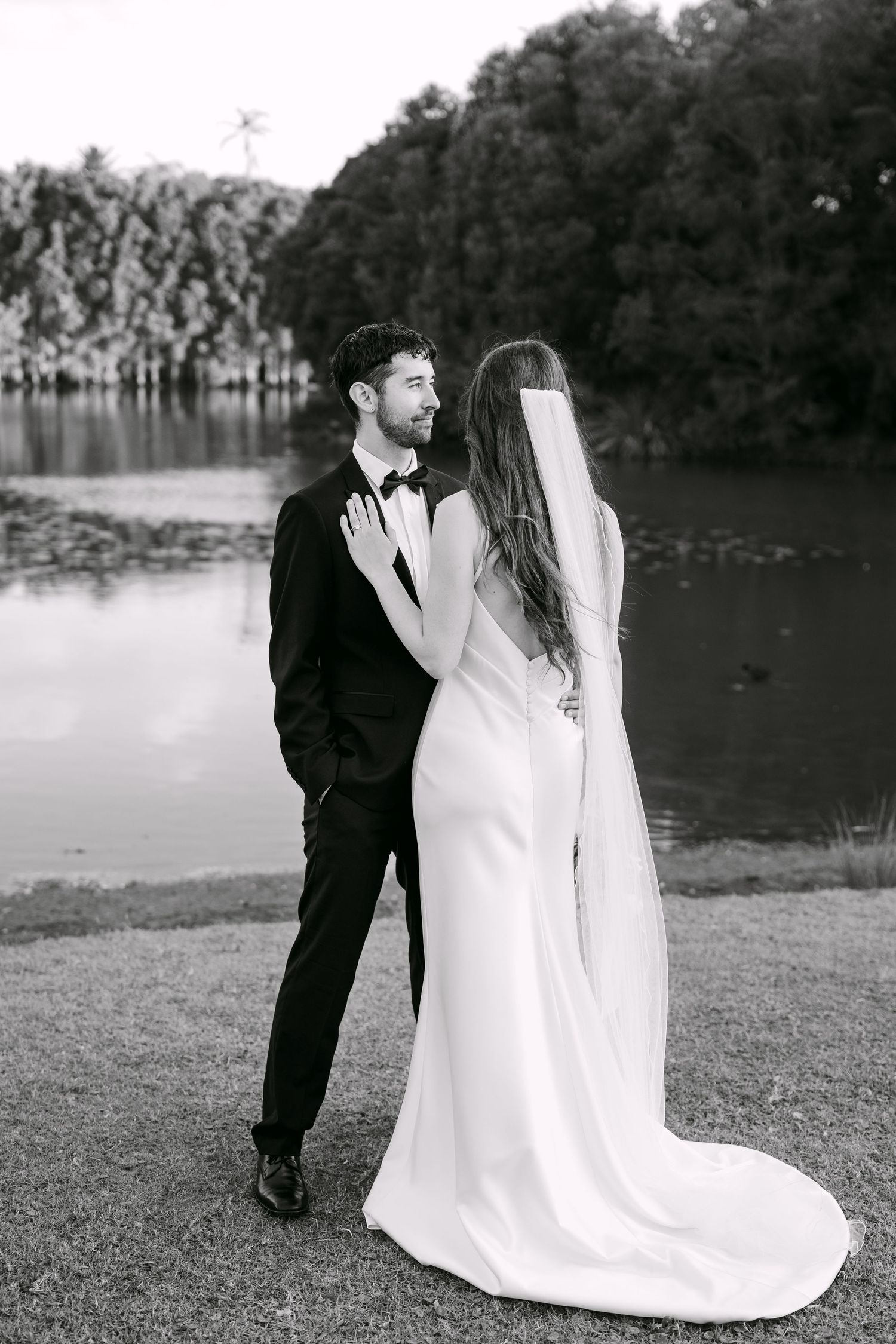 Black and white photo of a bride and groom standing by a lake, sharing a romantic moment in elegant wedding attire.