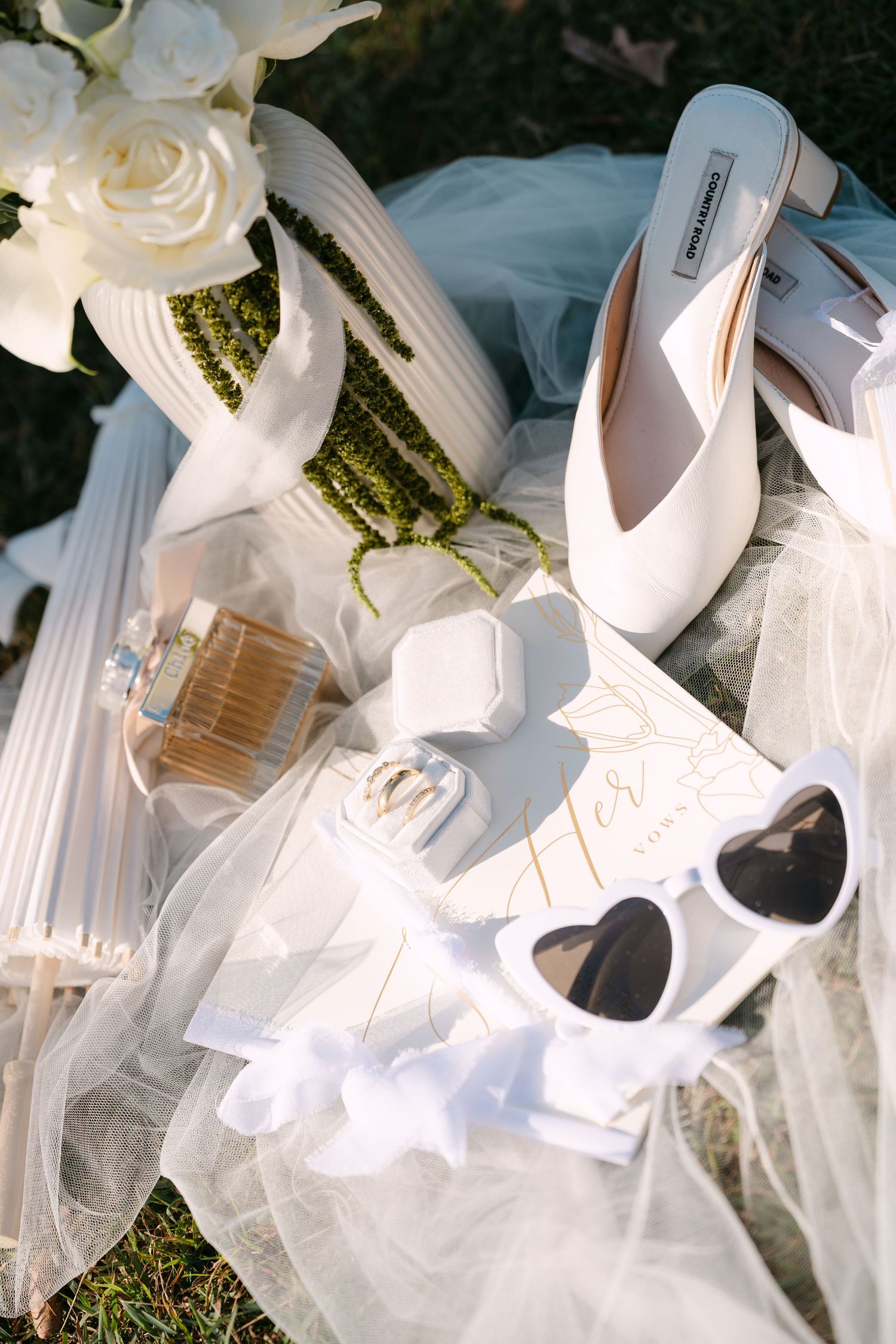 White bridal accessories flat lay featuring heels, heart sunglasses, perfume, jewelry box, and floral bouquet on tulle veil.