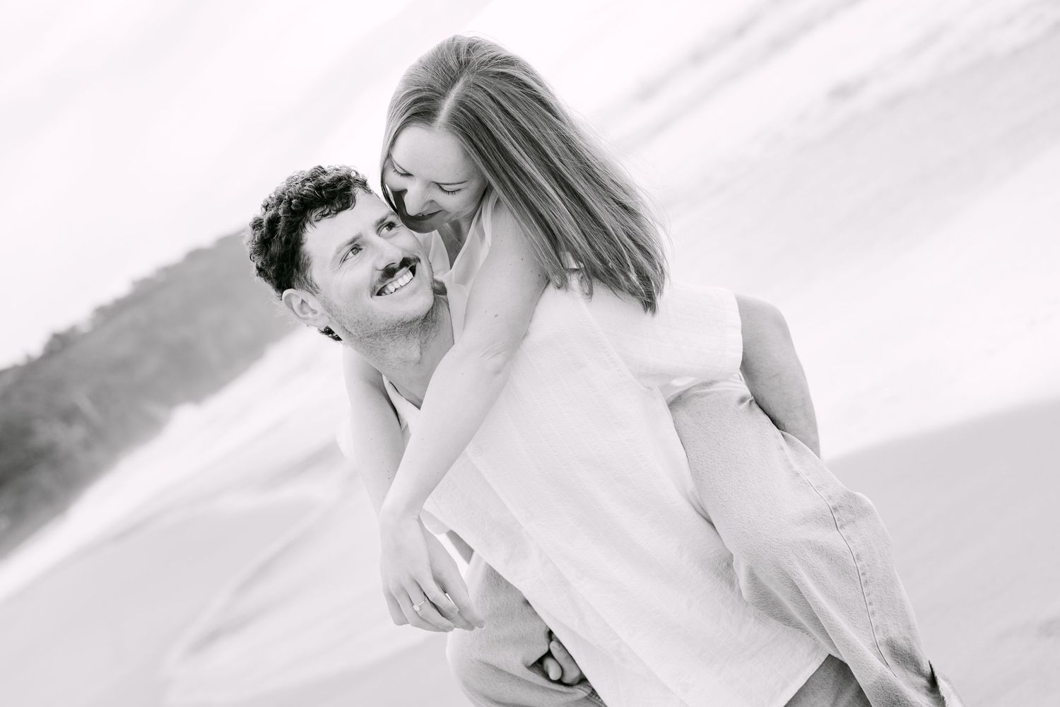 Black and white photo of laughing man giving woman a piggyback ride on a sunny beach.