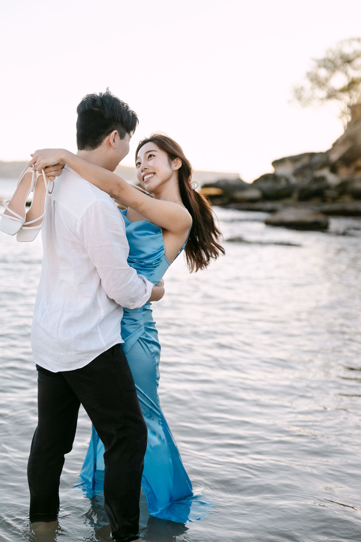 A couple embraces in shallow ocean water at sunset, the woman wearing a flowing blue gown.