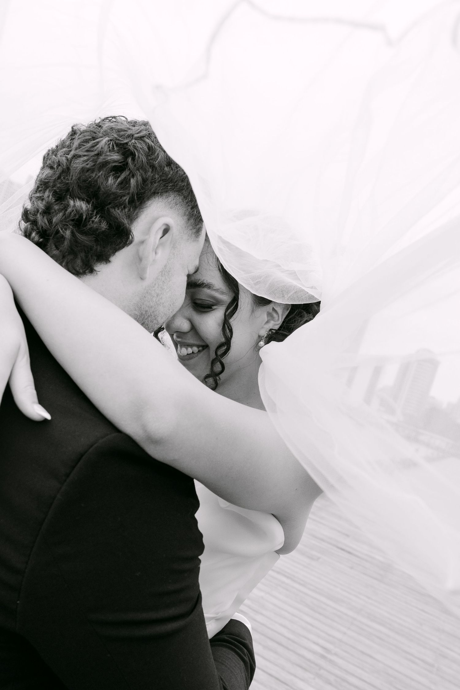 Black and white photo of a smiling bride and groom embracing under a flowing veil on their wedding day.