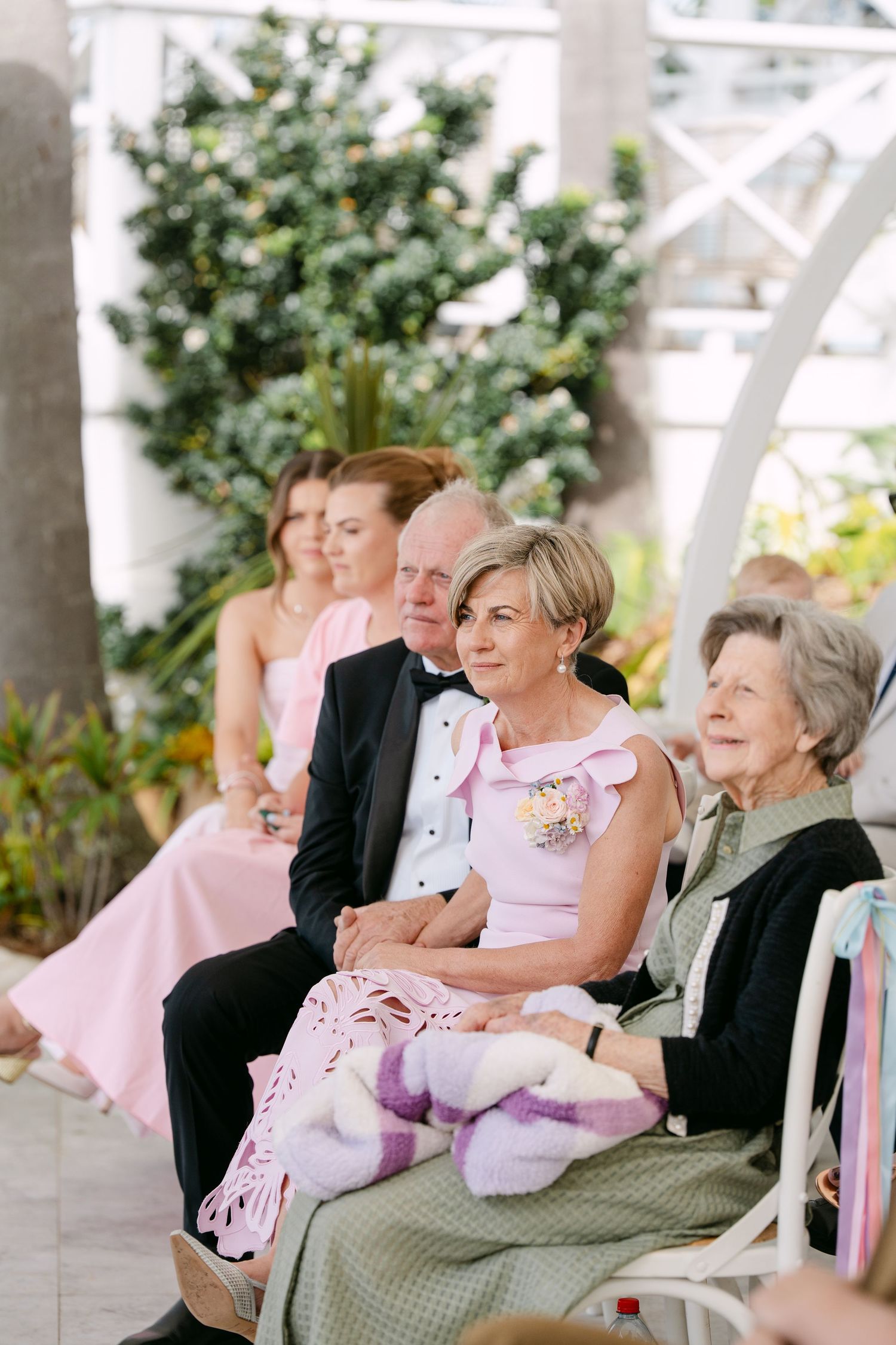 Elegant wedding guests in formal attire sit together on white chairs, watching a ceremony in a bright garden setting with lush greenery.