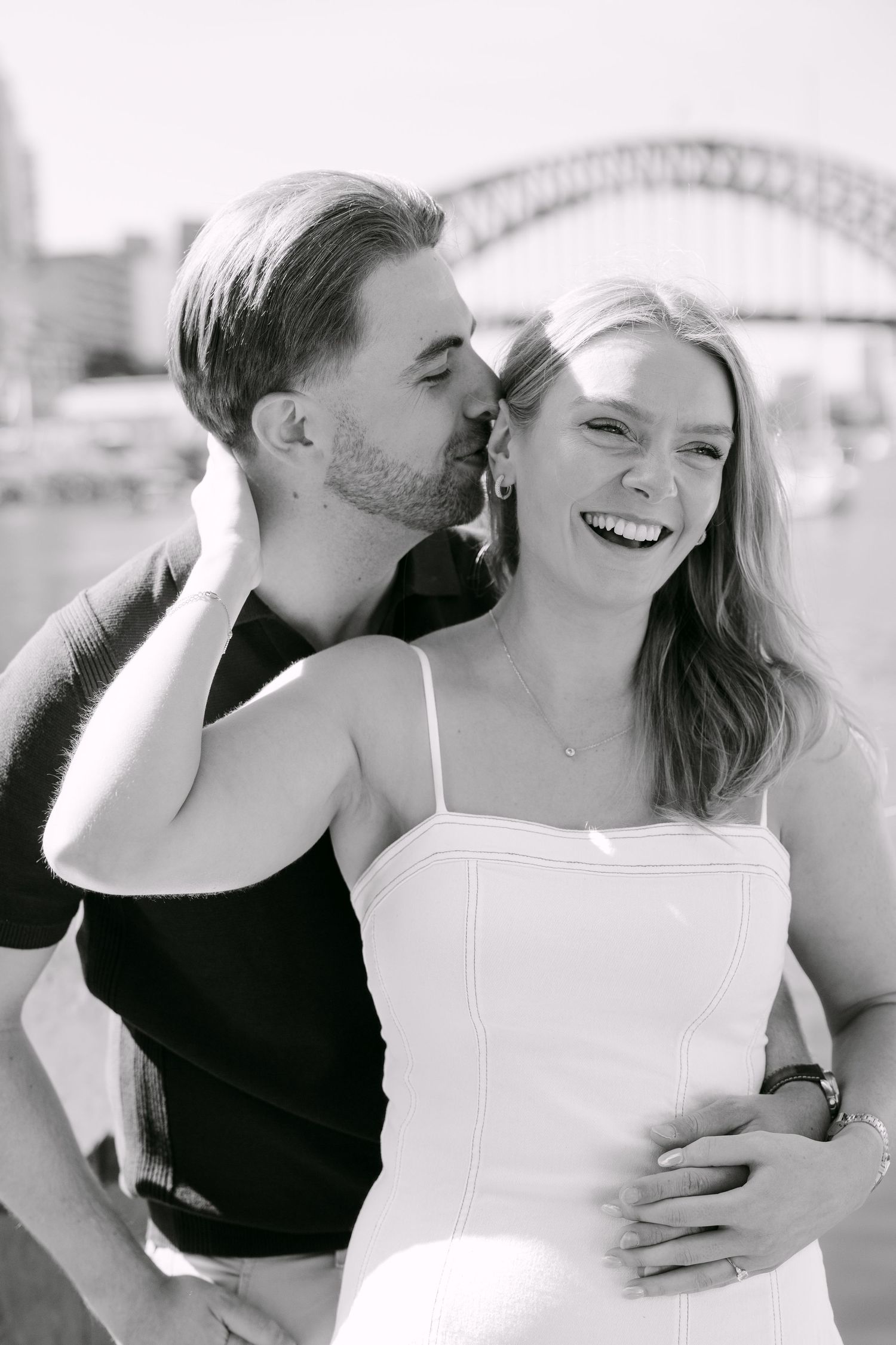 Black and white photo of a couple embracing near Sydney Harbour Bridge, man kissing woman's cheek as she laughs.