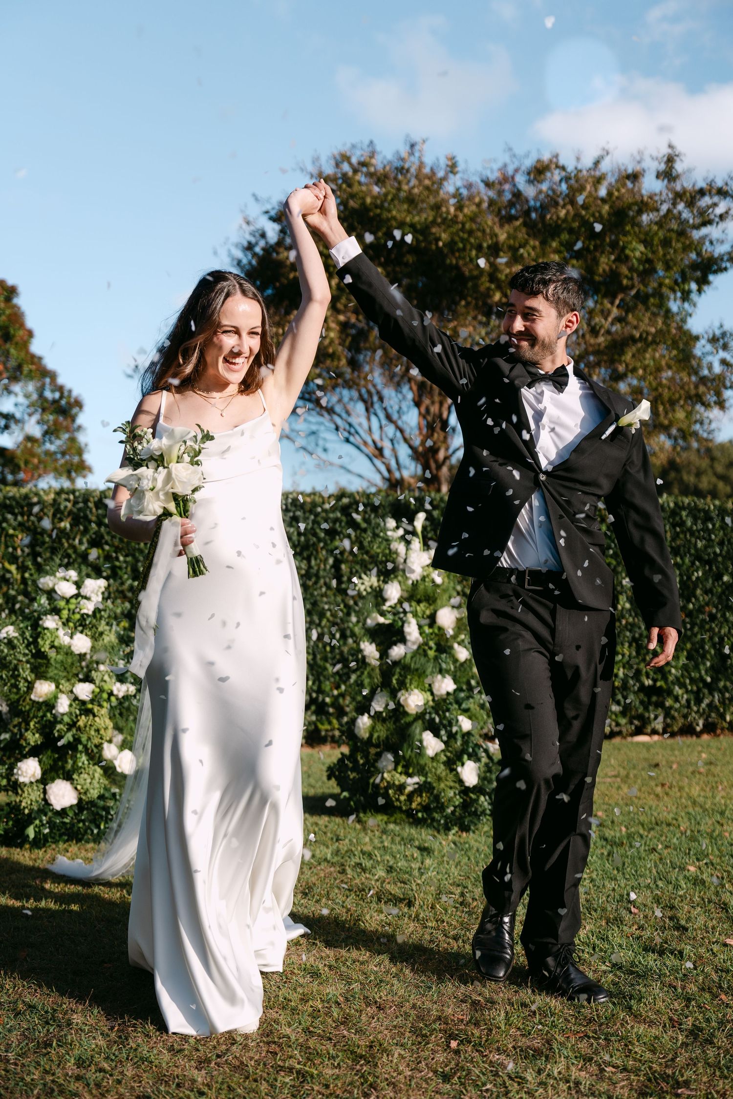 Joyful bride and groom celebrating outdoors, raising hands with confetti, white floral backdrop and blue sky.