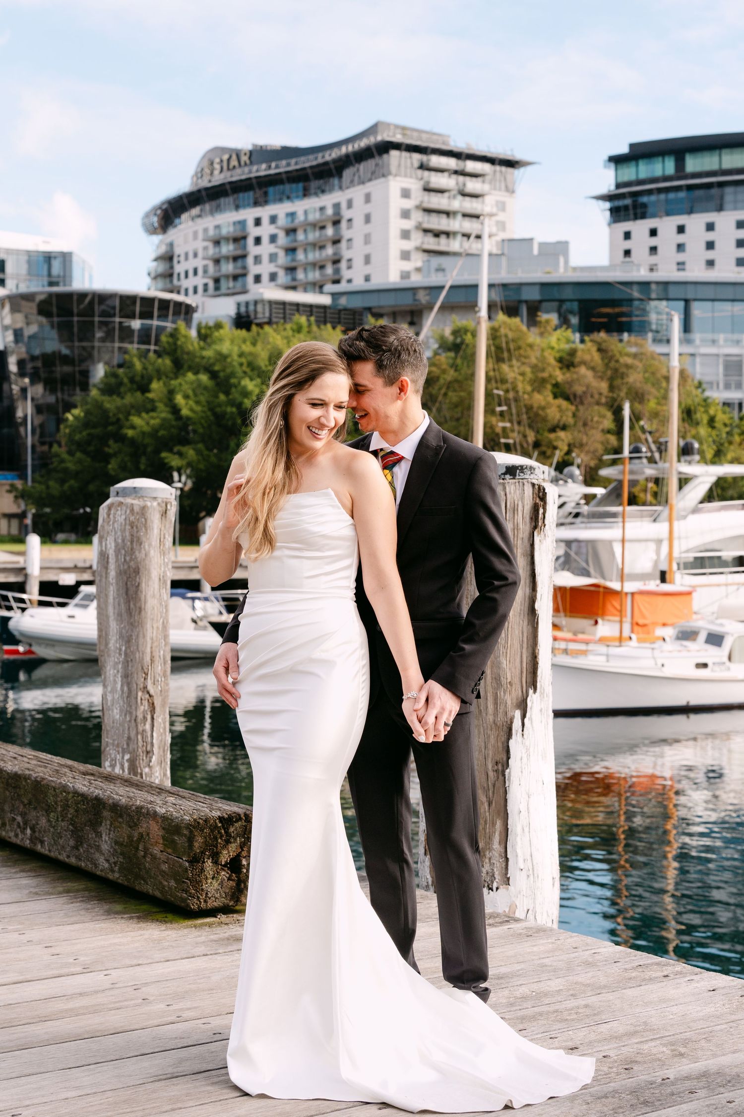 Bride and groom holding hands on a waterfront dock with marina and modern buildings in the background.