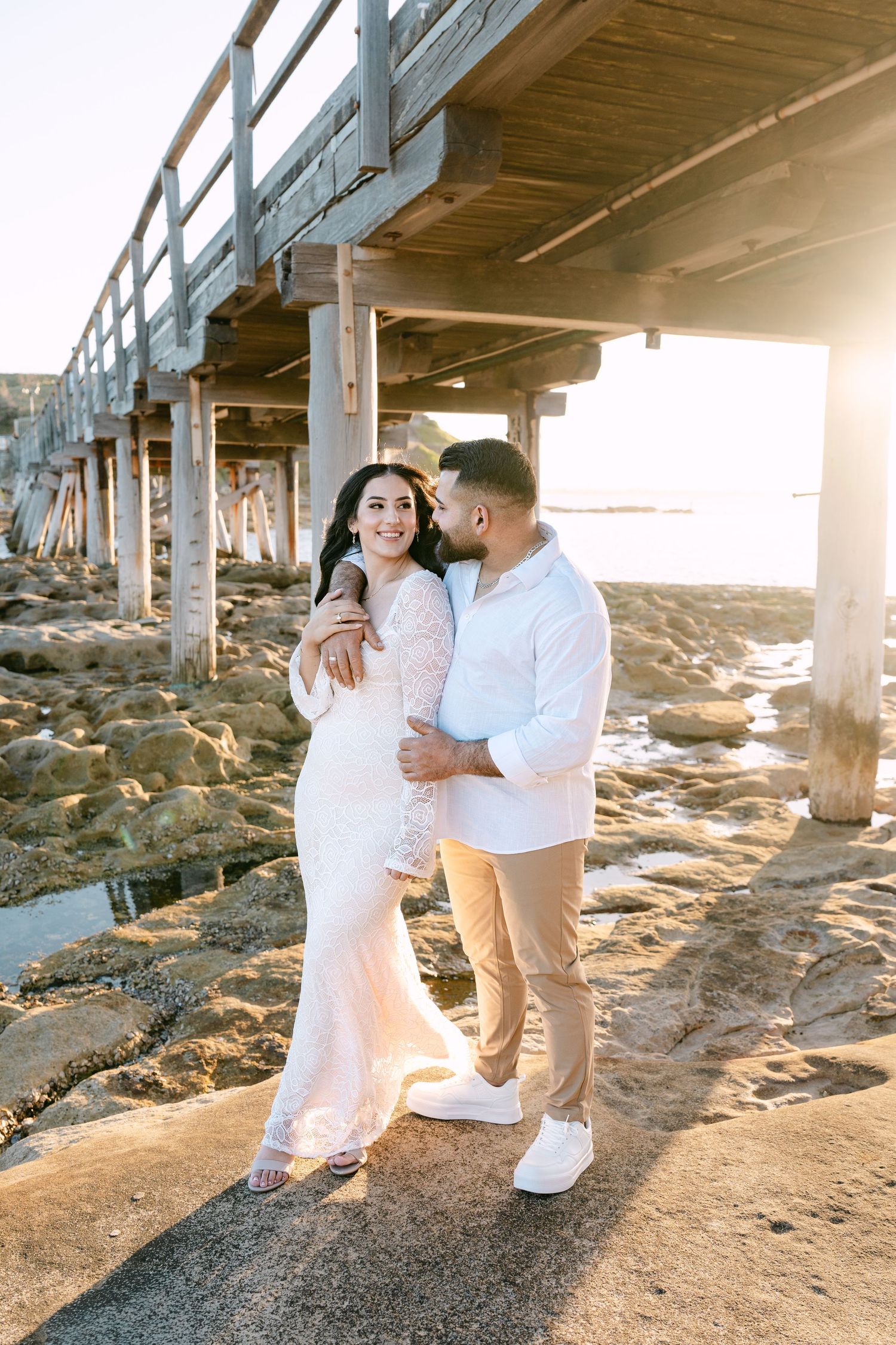 Romantic couple holding each other under a pier on rocky shore, woman in white lace gown smiling.
