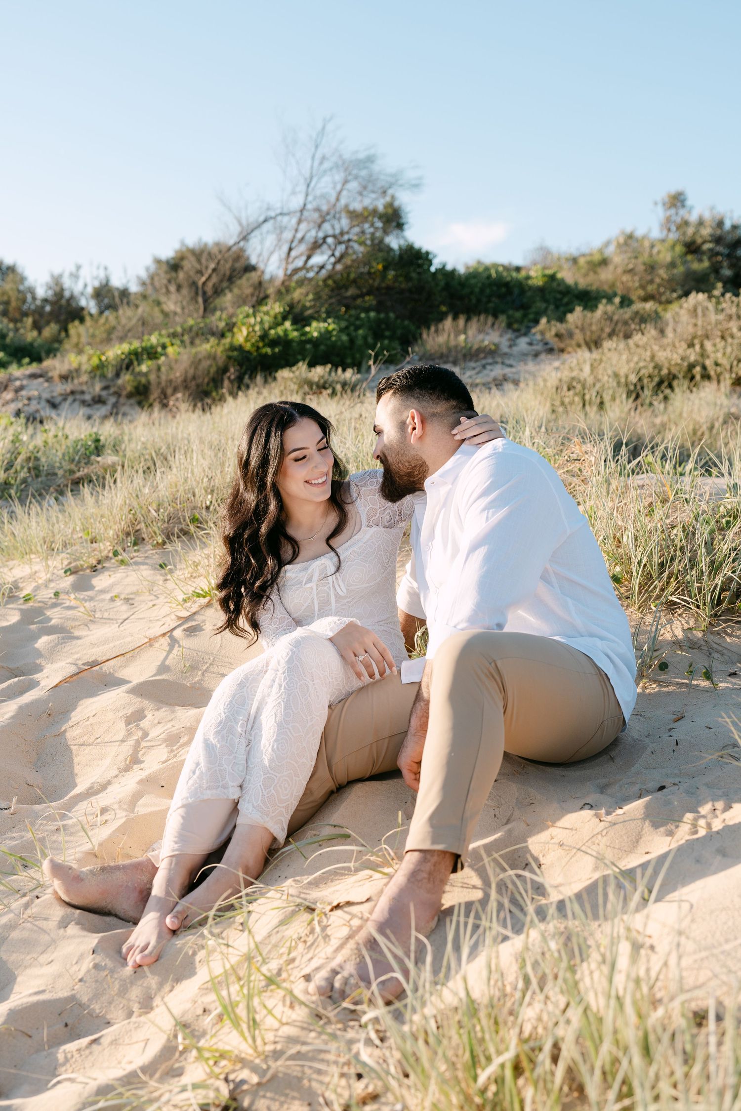 Couple sitting together on sandy dunes, sharing a tender moment under a clear blue sky.