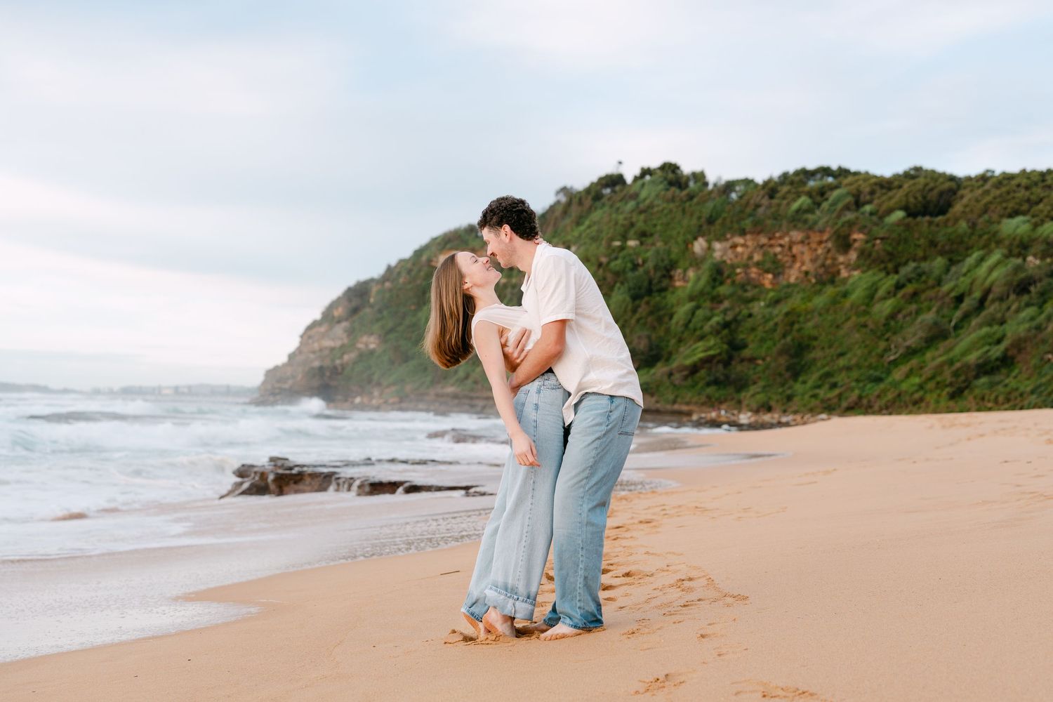Romantic couple sharing a kiss on sandy beach with lush green headland behind.