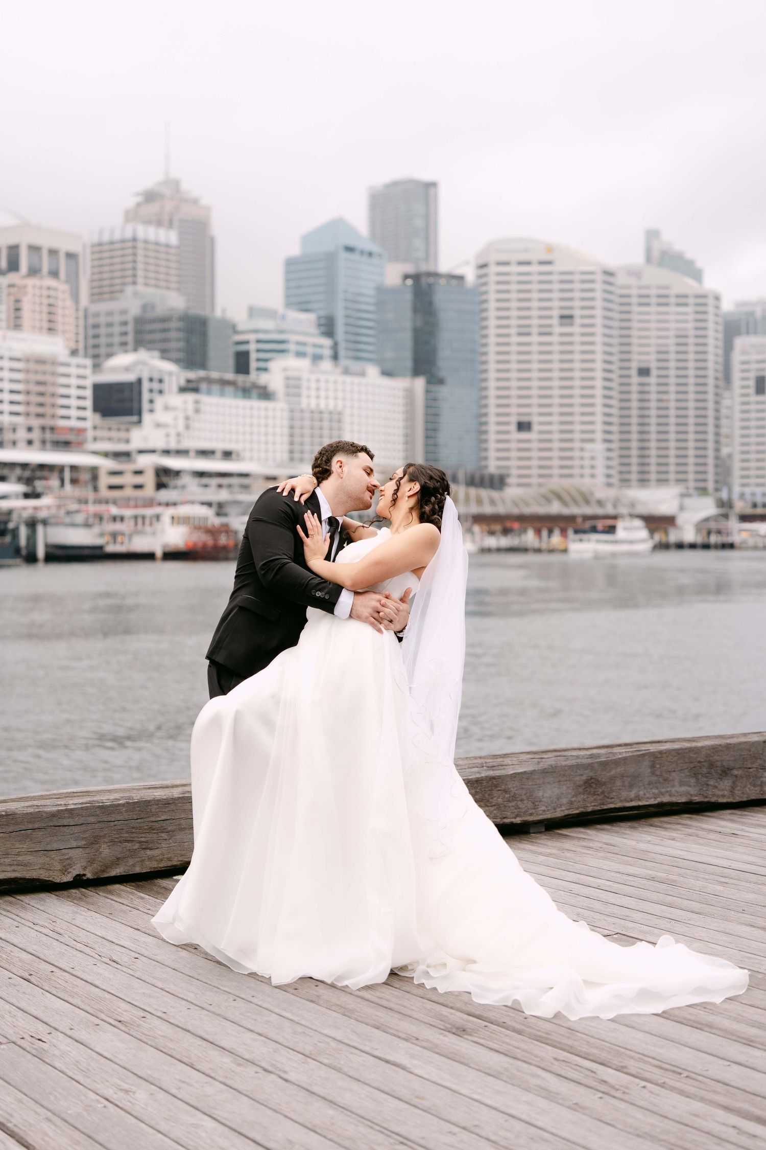Bride and groom kissing on a waterfront pier with a city skyline backdrop on their wedding day.