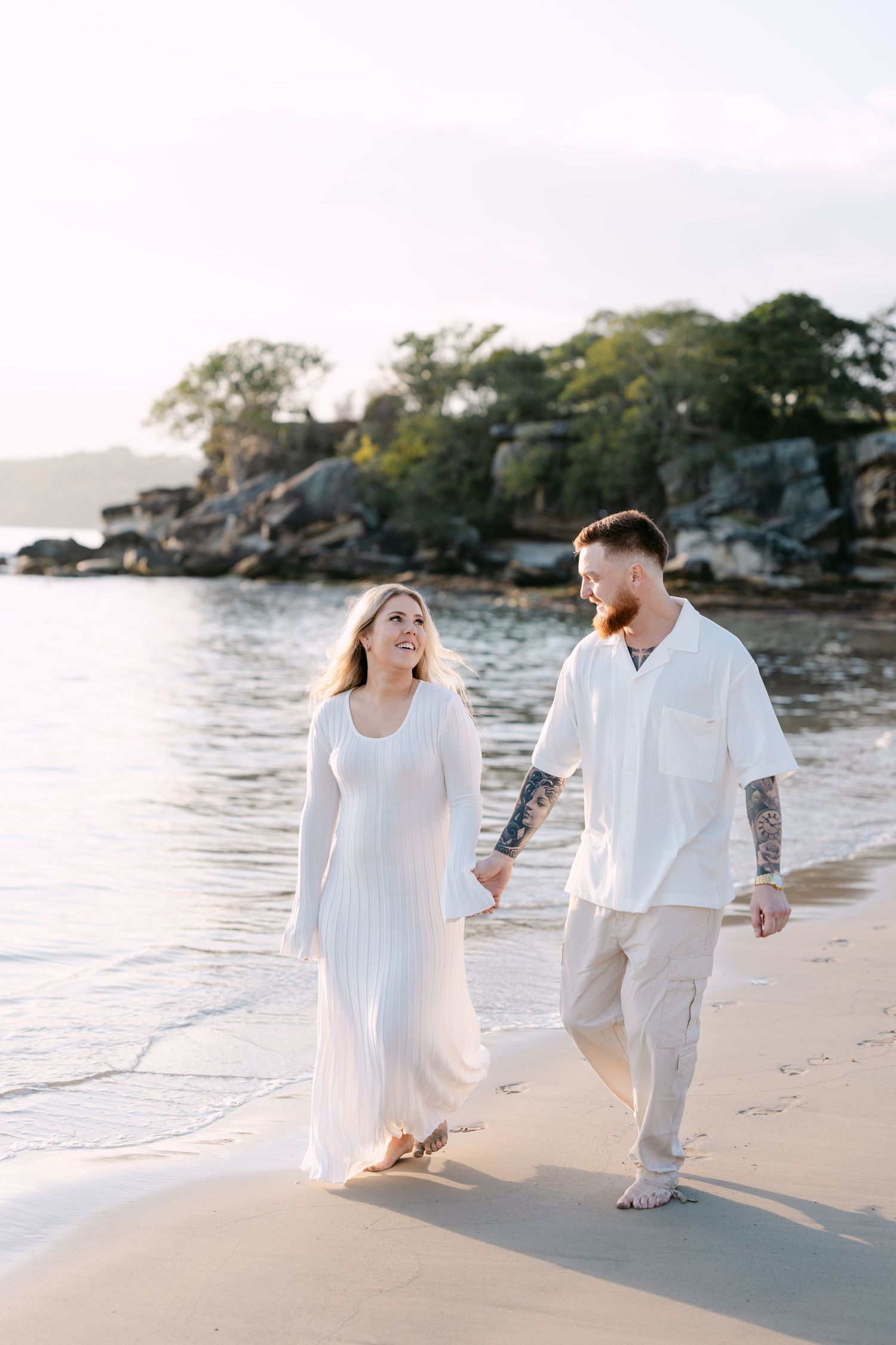 Couple in white outfits holding hands and walking barefoot on a sandy beach at golden hour.