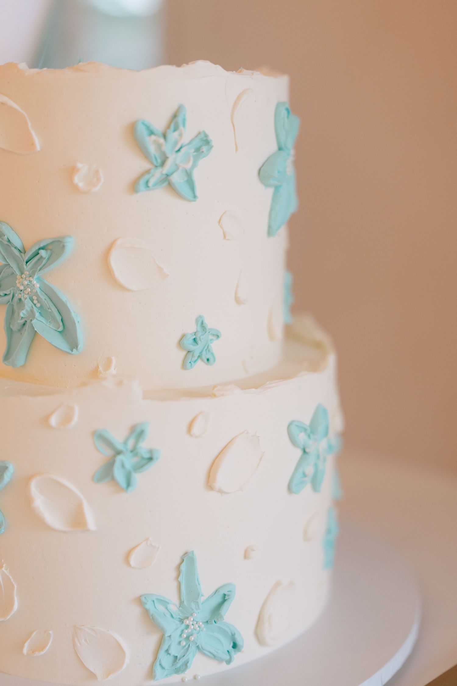 Two-tier white buttercream wedding cake decorated with hand-painted teal starfish and shell accents on a white cake stand.
