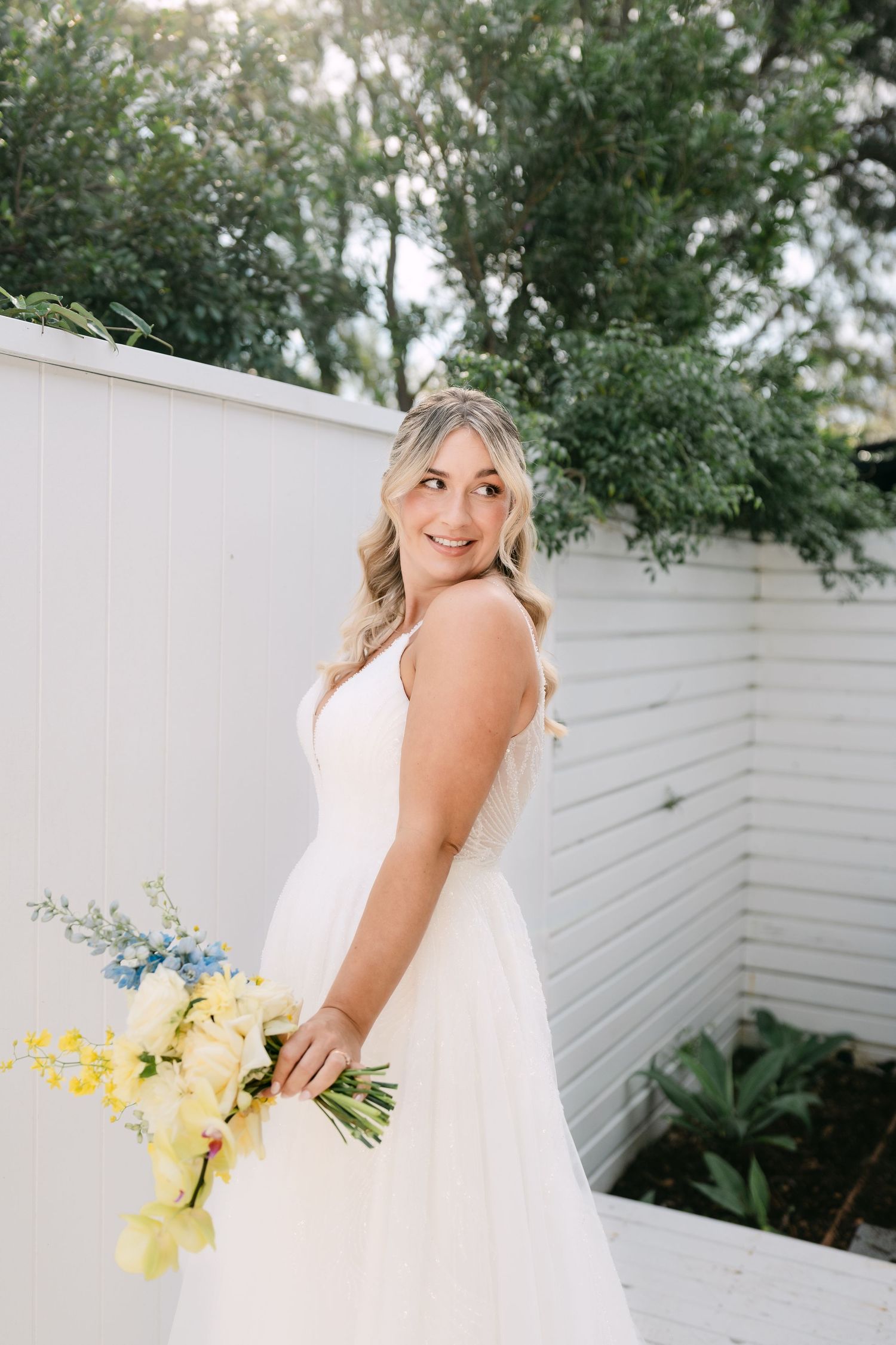 Smiling bride in a white tulle wedding gown holds a yellow and blue floral bouquet while glancing over her shoulder in a sunny garden.