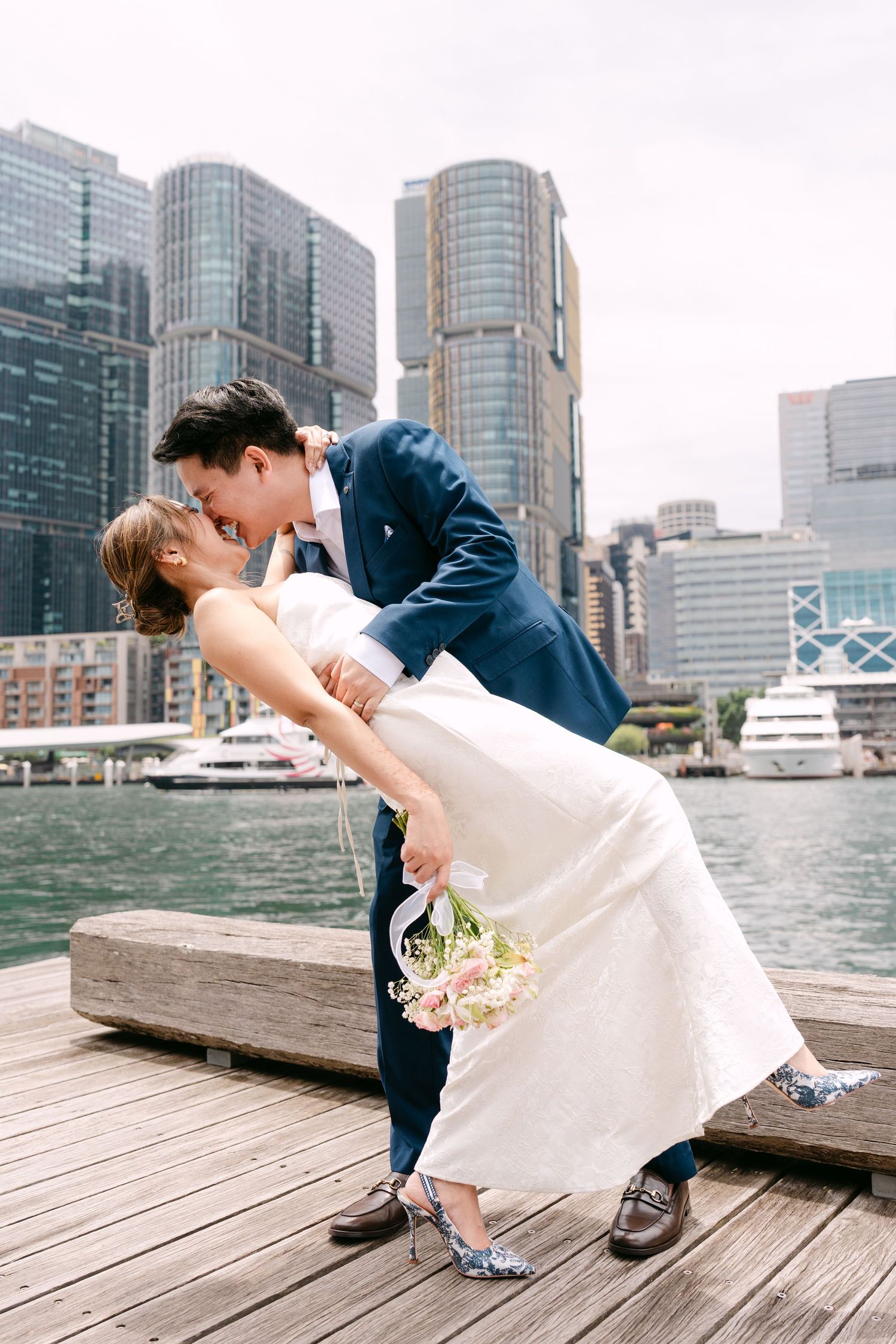 Groom dipping bride for a kiss on a harbor pier with city skyline in the background.