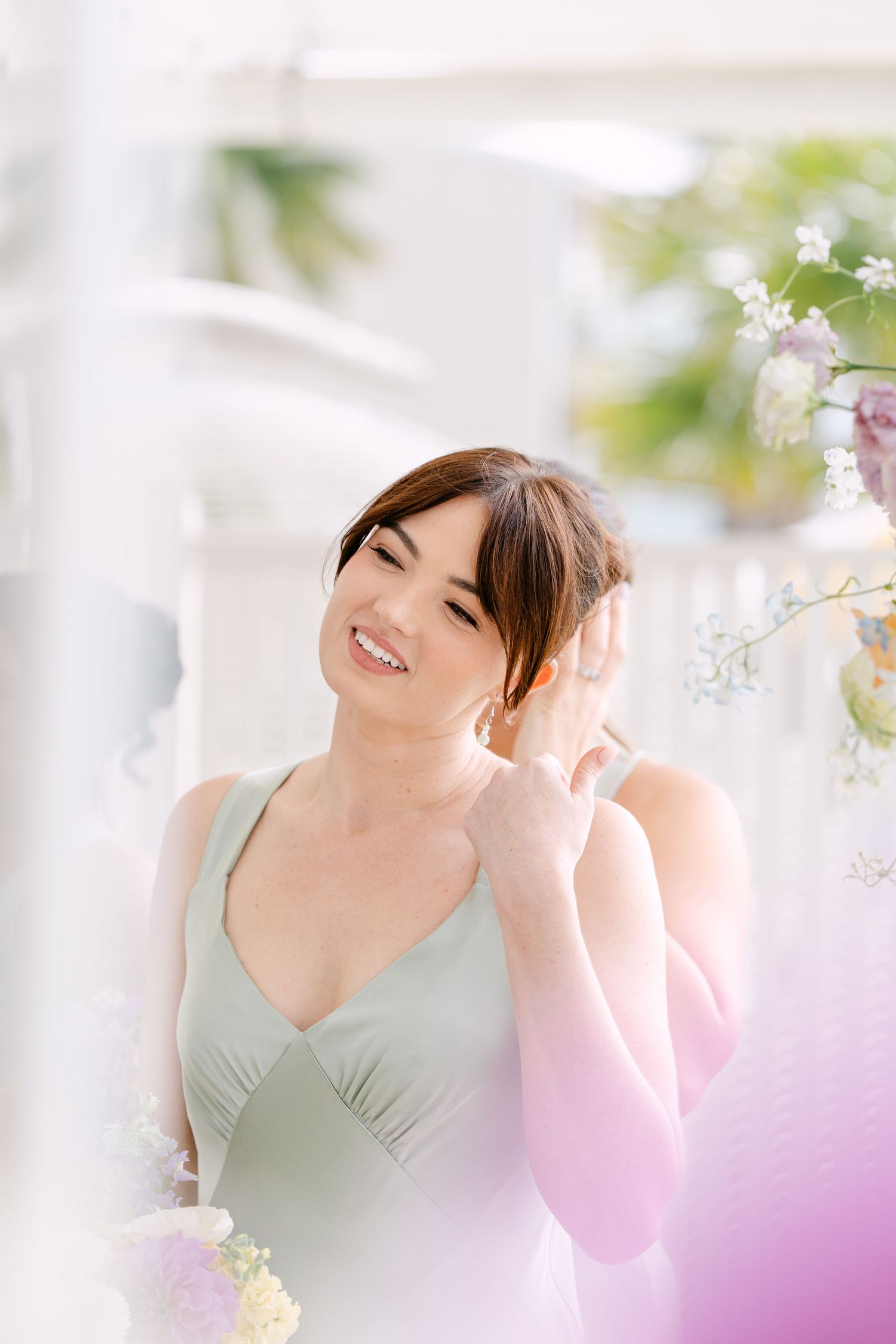 Smiling bridesmaid in a sage green satin dress touches her hair near delicate pastel wedding flowers.