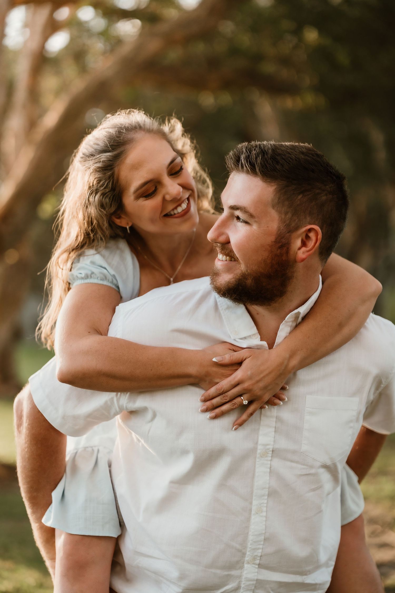 Woman in blue dress hugging man from behind piggyback style laughing in a sunny park setting.