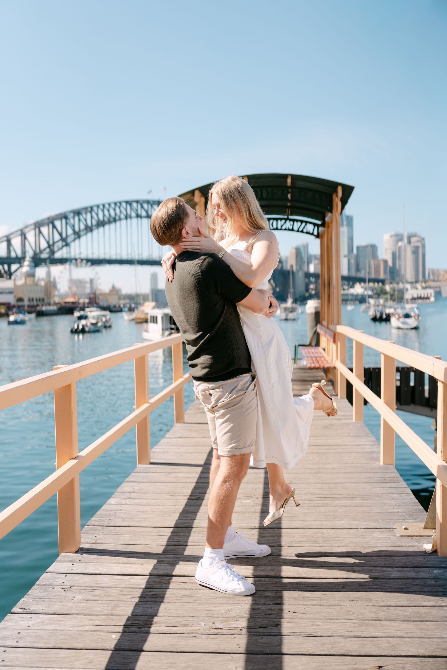Man lifting woman for a kiss on a wooden pier with Sydney Harbour Bridge behind them.