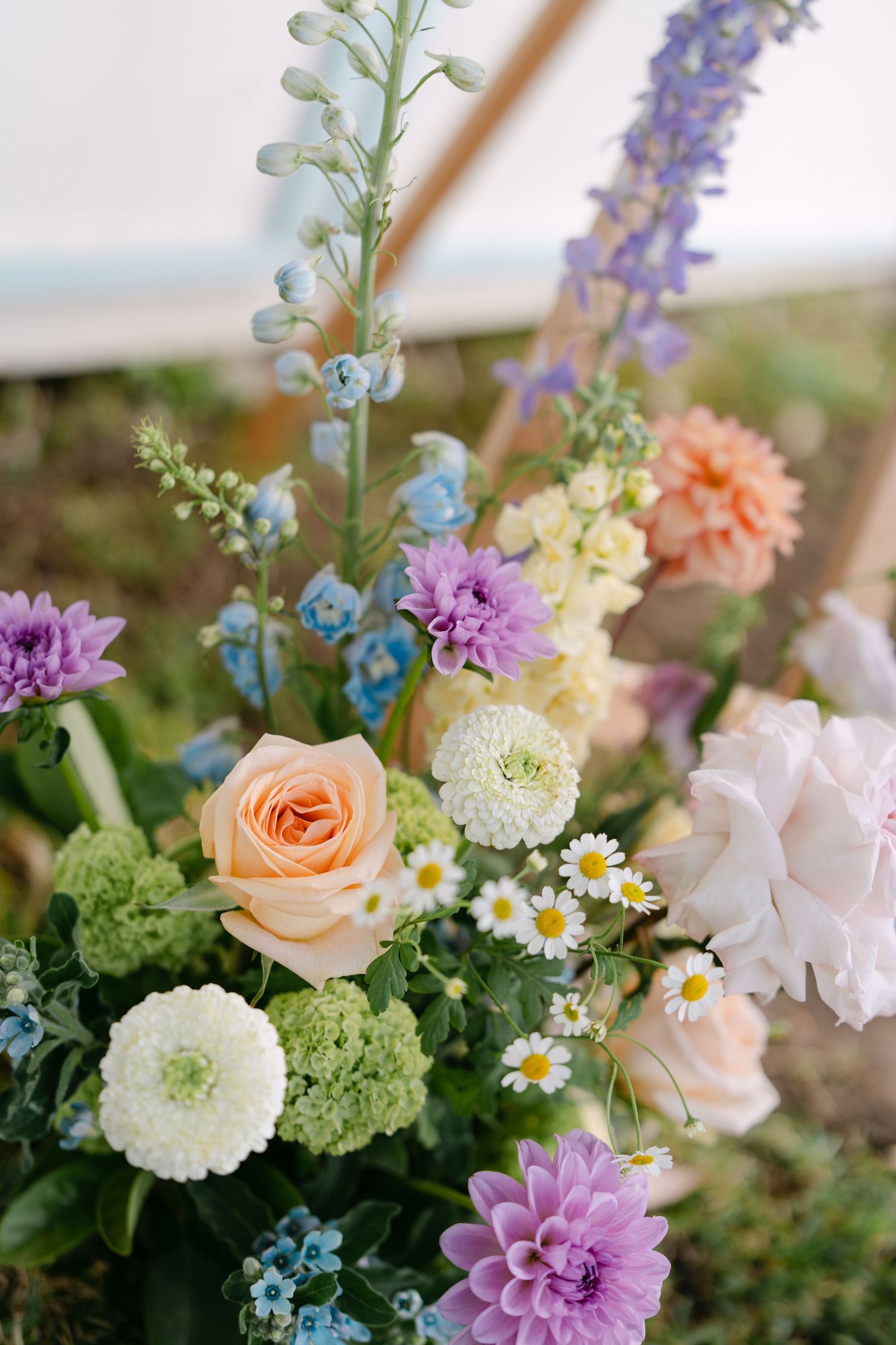Pastel wedding flower arrangement with peach roses, purple dahlias, blue delphinium, white zinnias and daisies, and green hydrangea.