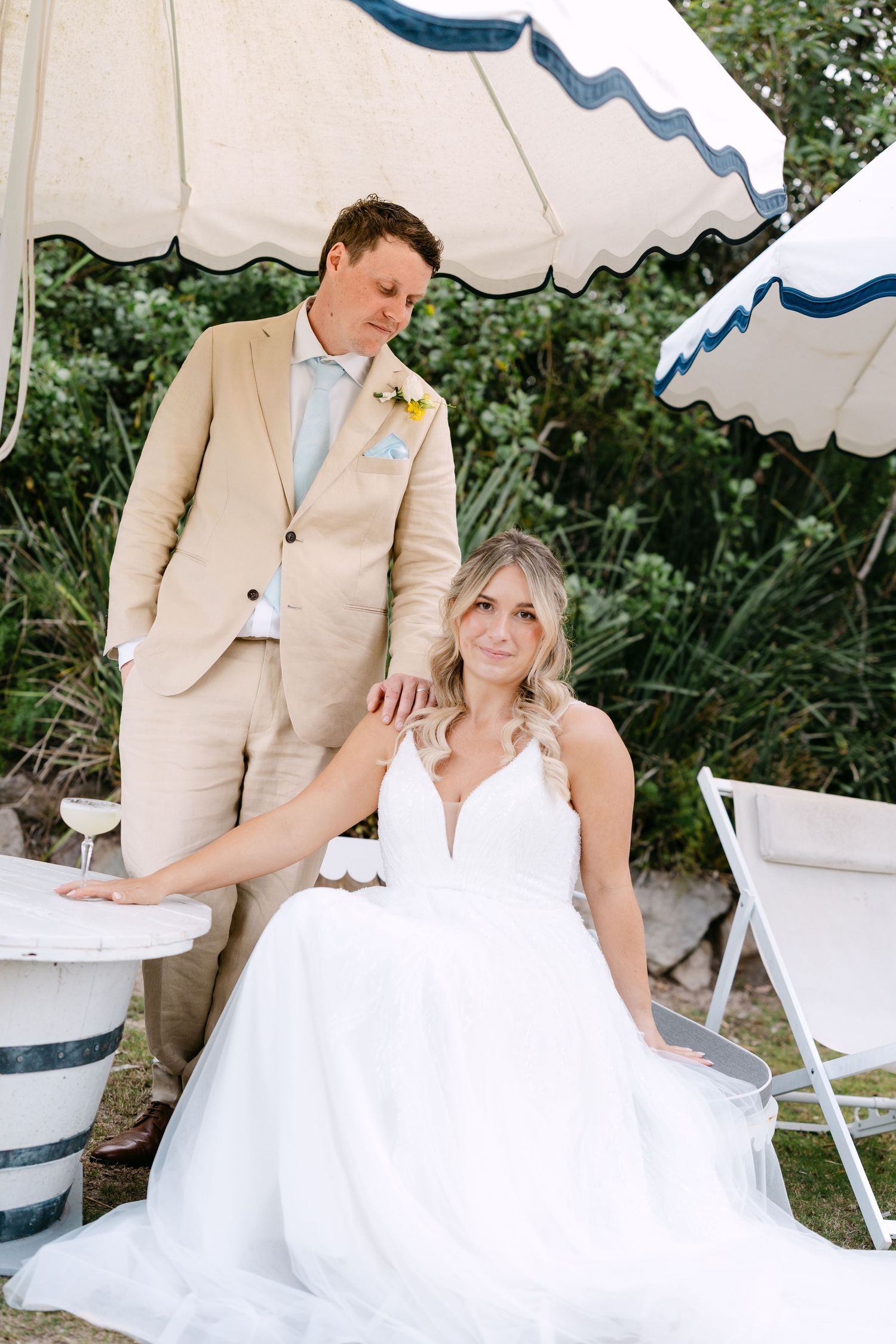 Editorial Style photo of Bride in white gown sits while groom in tan suit stands under striped beach umbrella.