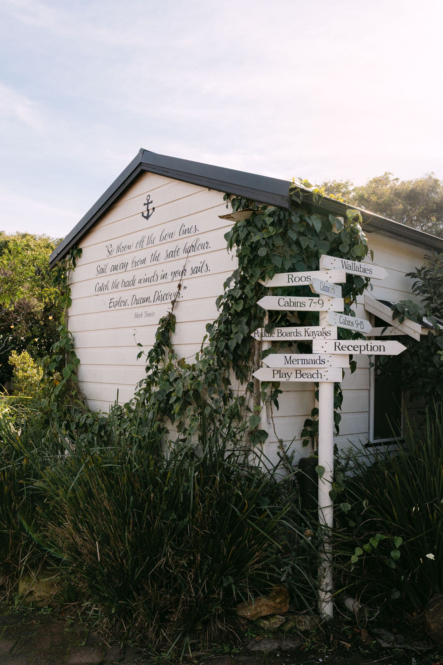 White coastal building with ivy, anchor motif, inspirational quote, and directional signpost pointing to cabins, reception, and beach.