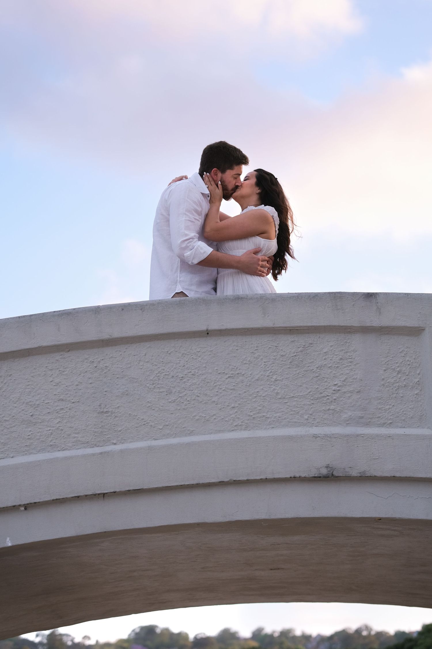 Couple kissing on top of a white arched bridge against a pastel sunset sky.