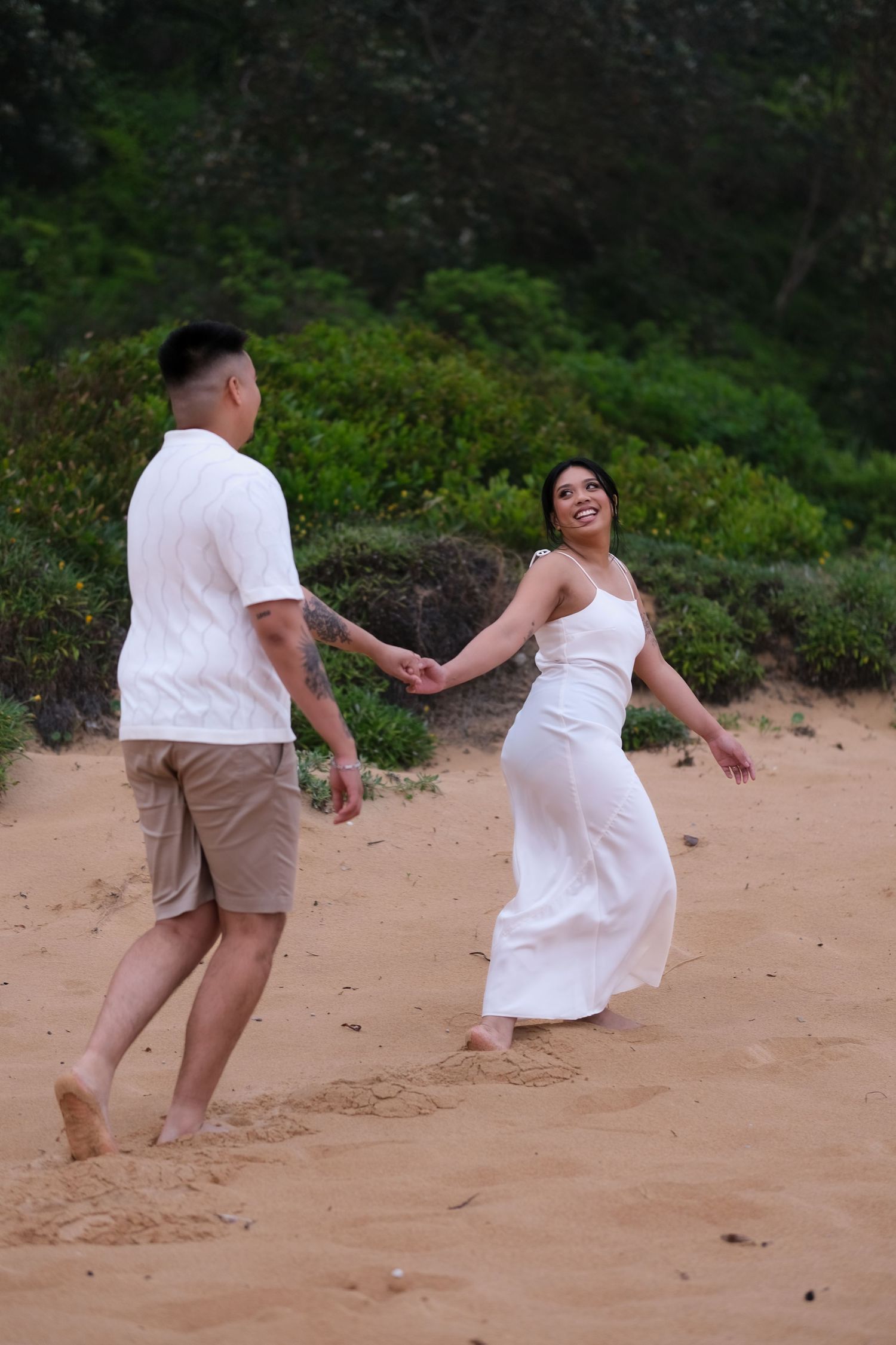 Couple holding hands and dancing playfully on a sandy beach with green shrubs behind.