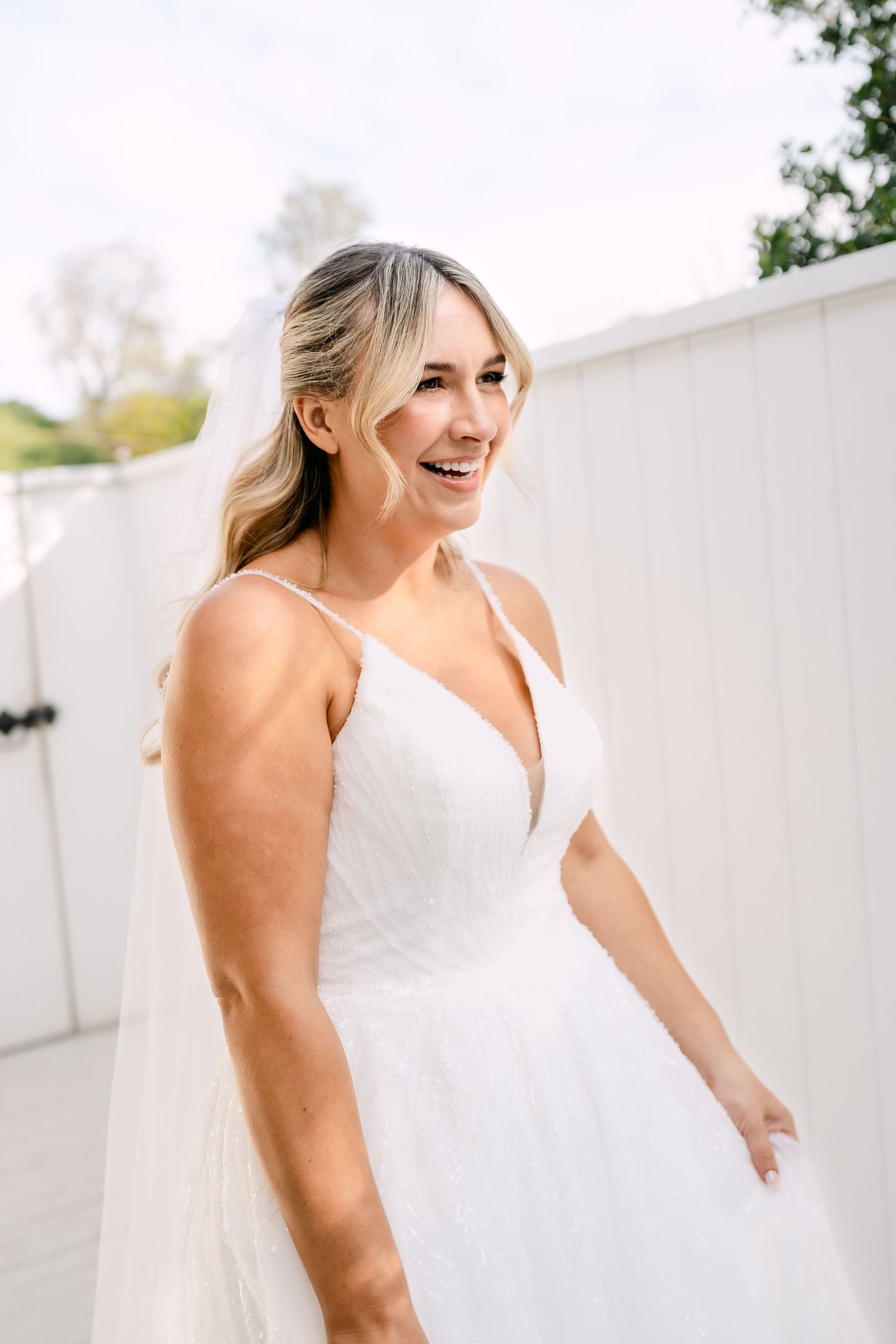 Smiling bride in a white beaded V-neck wedding gown and long veil stands outdoors beside a white fence on a bright, sunny wedding day.