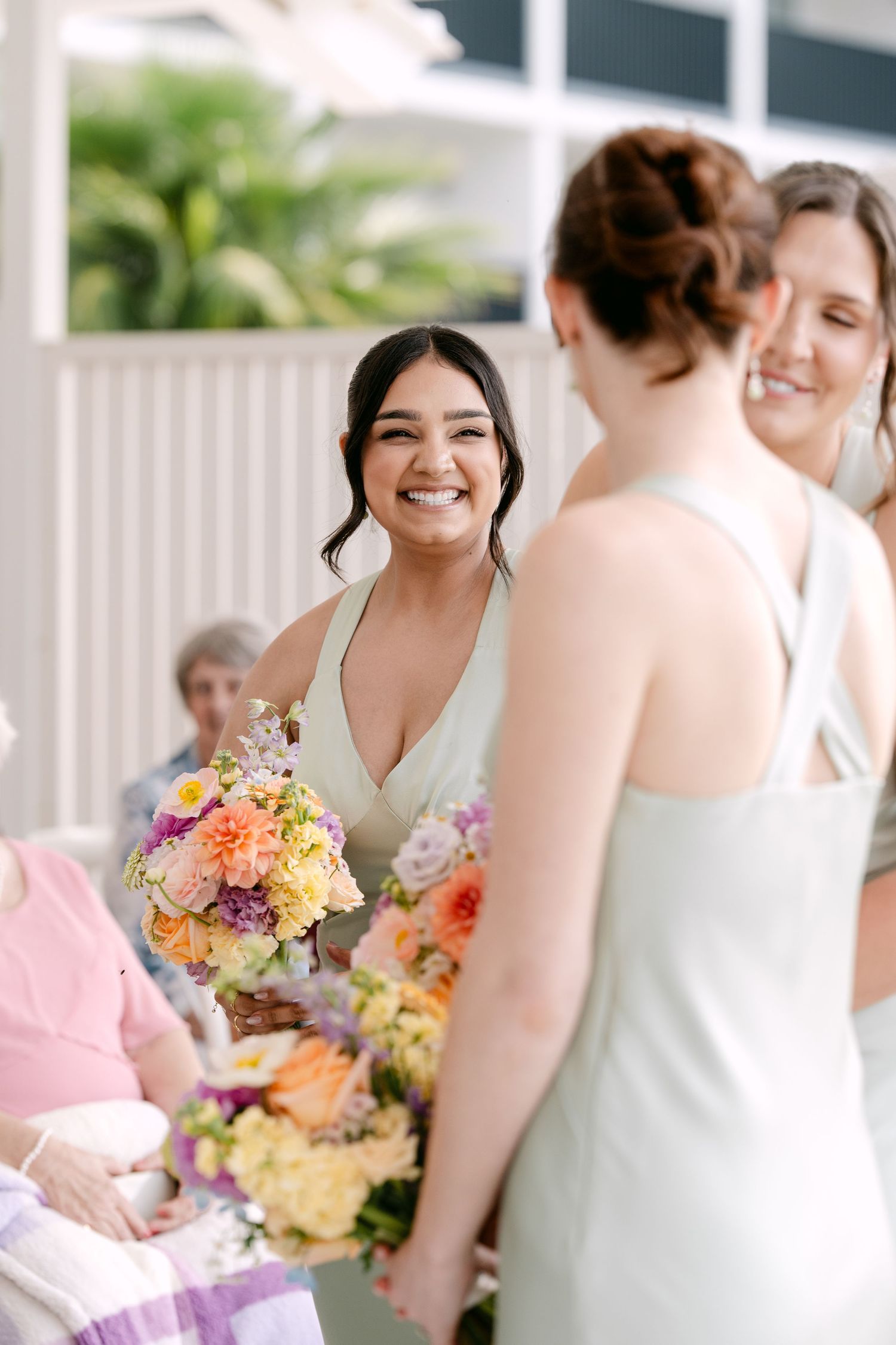 Smiling bridesmaid in a sage green dress holds a colorful peach and lavender bouquet while chatting with other bridesmaids at a wedding.