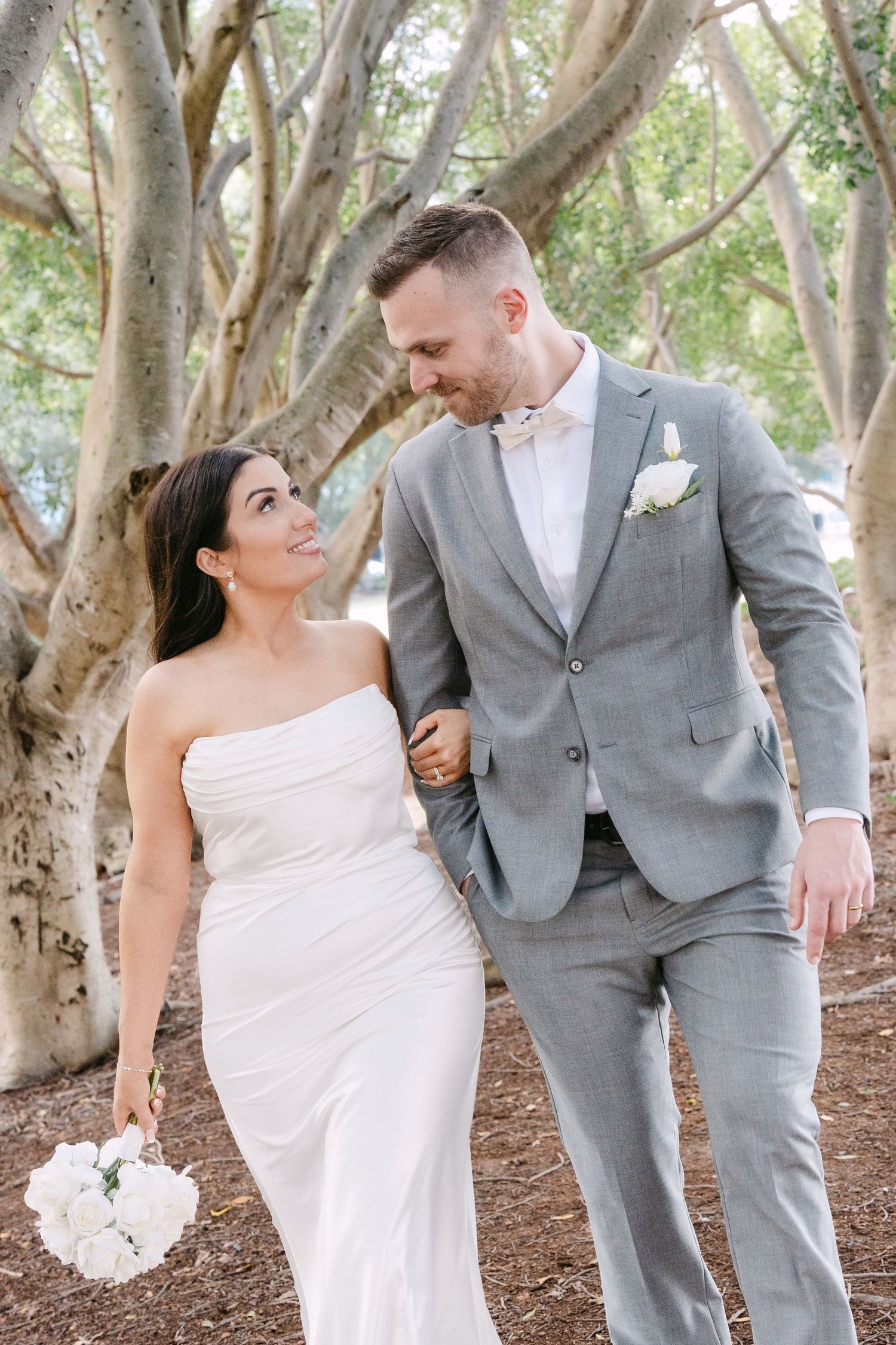 Smiling bride in a white strapless gown holding white roses walks arm in arm with a groom in a gray suit under large trees.