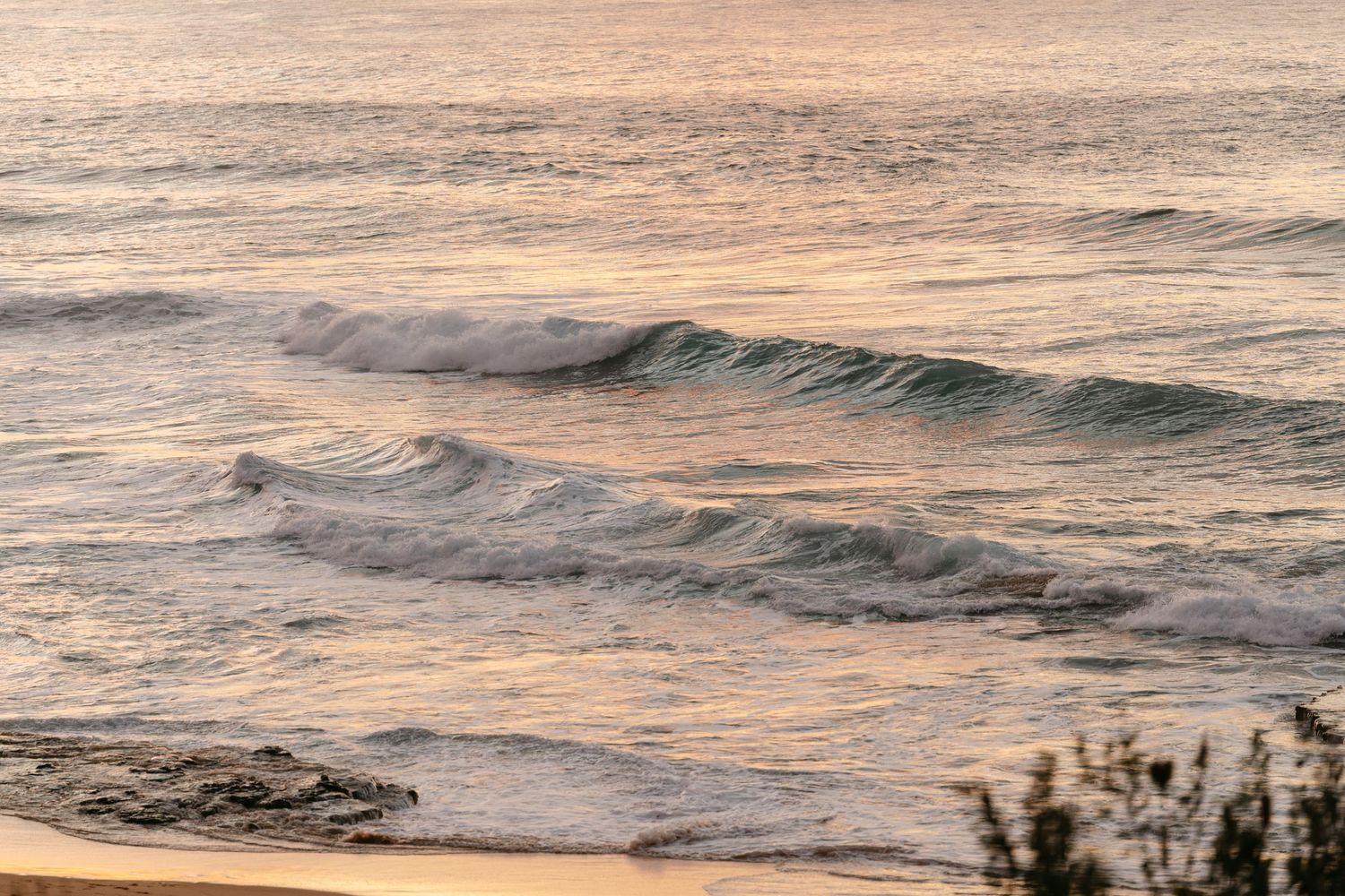 Golden hour ocean waves crashing on a sandy beach at sunset with soft pastel light reflecting on the water.
