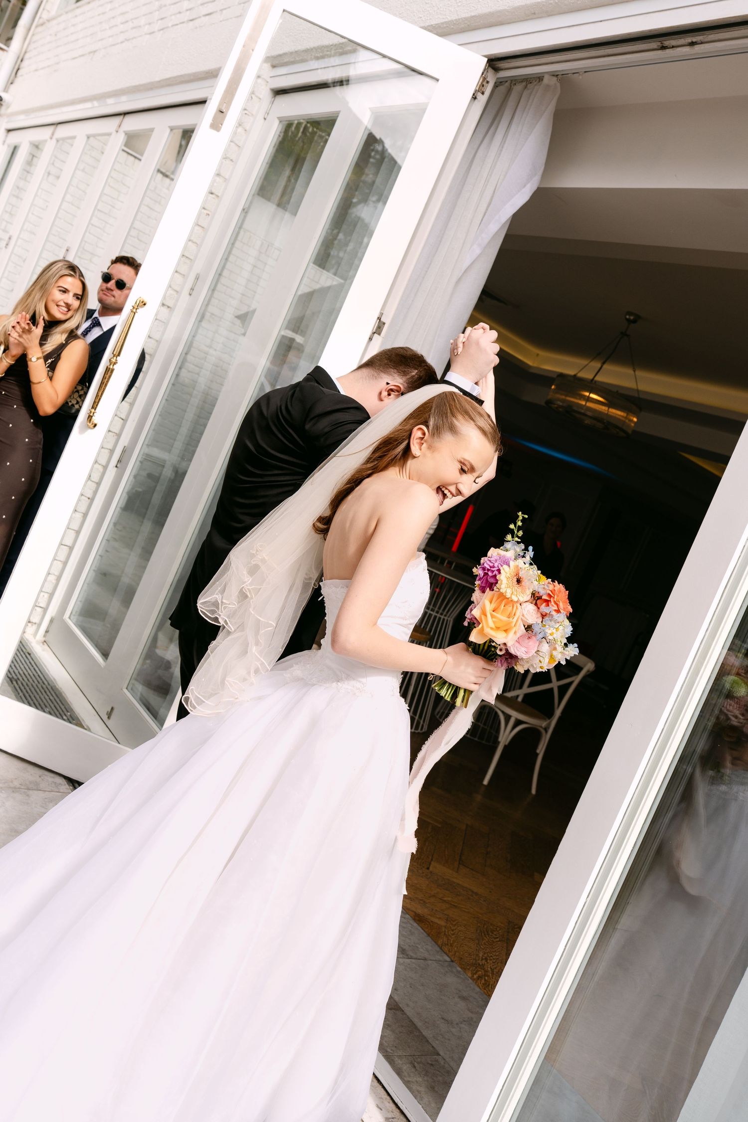 Smiling bride in a white strapless gown holding a colorful bouquet as she and the groom walk through glass doors while guests applaud.