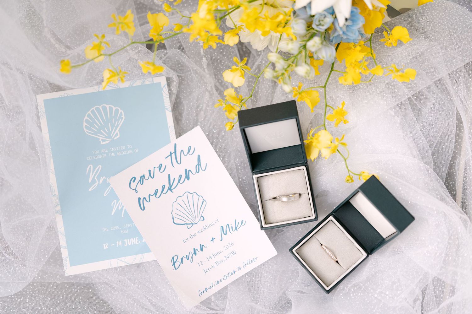 Wedding save the date cards with seashell design beside two ring boxes with bands.