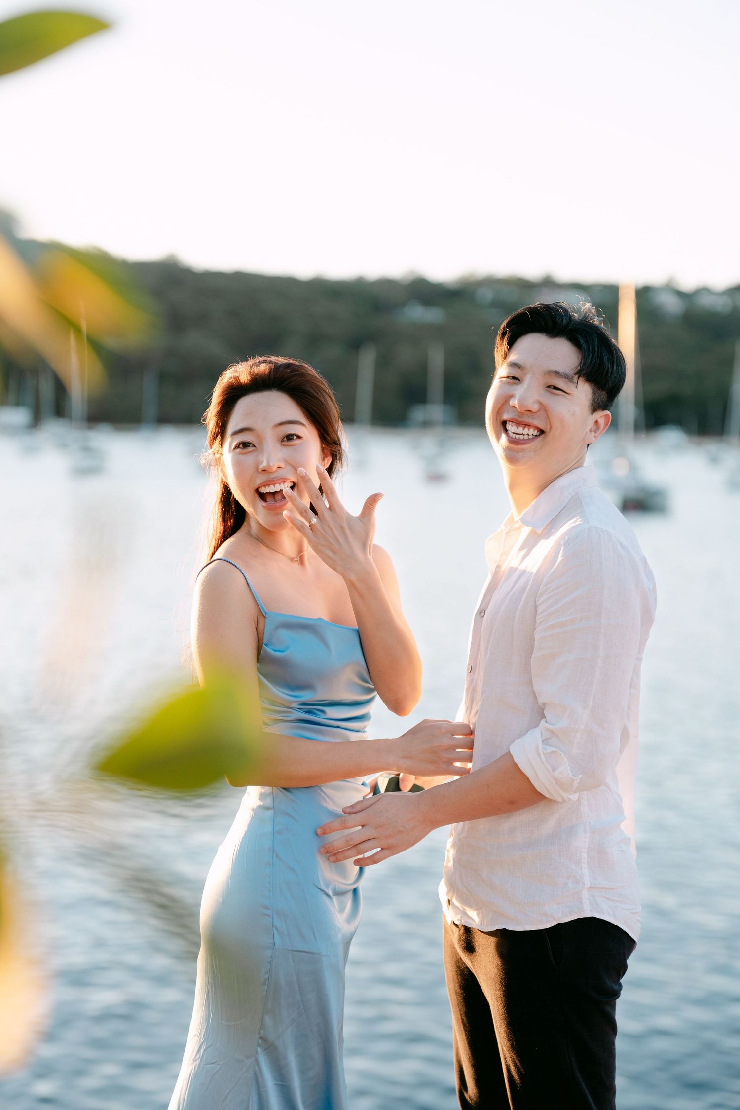 A smiling couple celebrates their engagement by a waterfront at golden hour, the woman showing her ring.