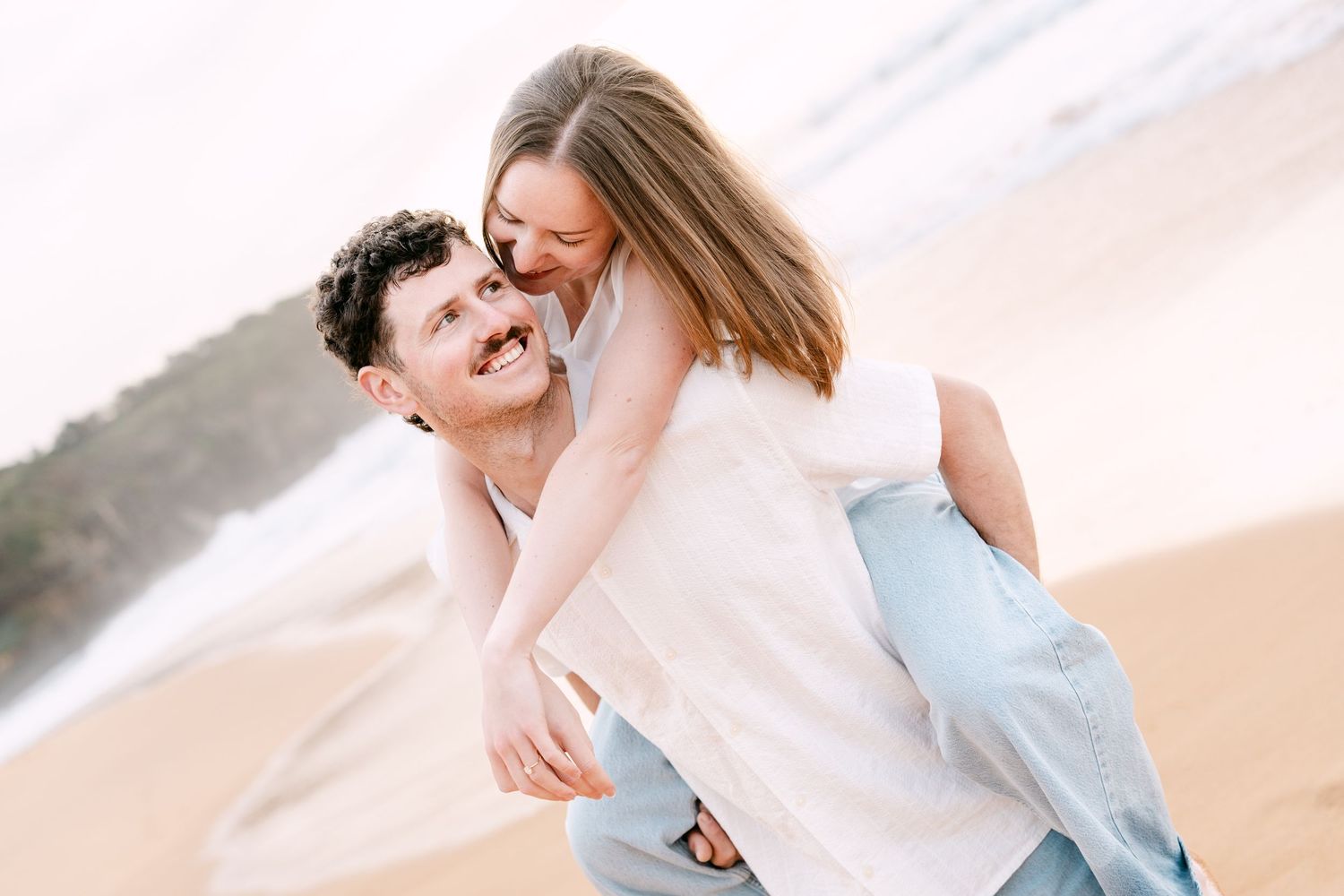 Couple laughing on beach, man giving woman piggyback ride on sandy shore.