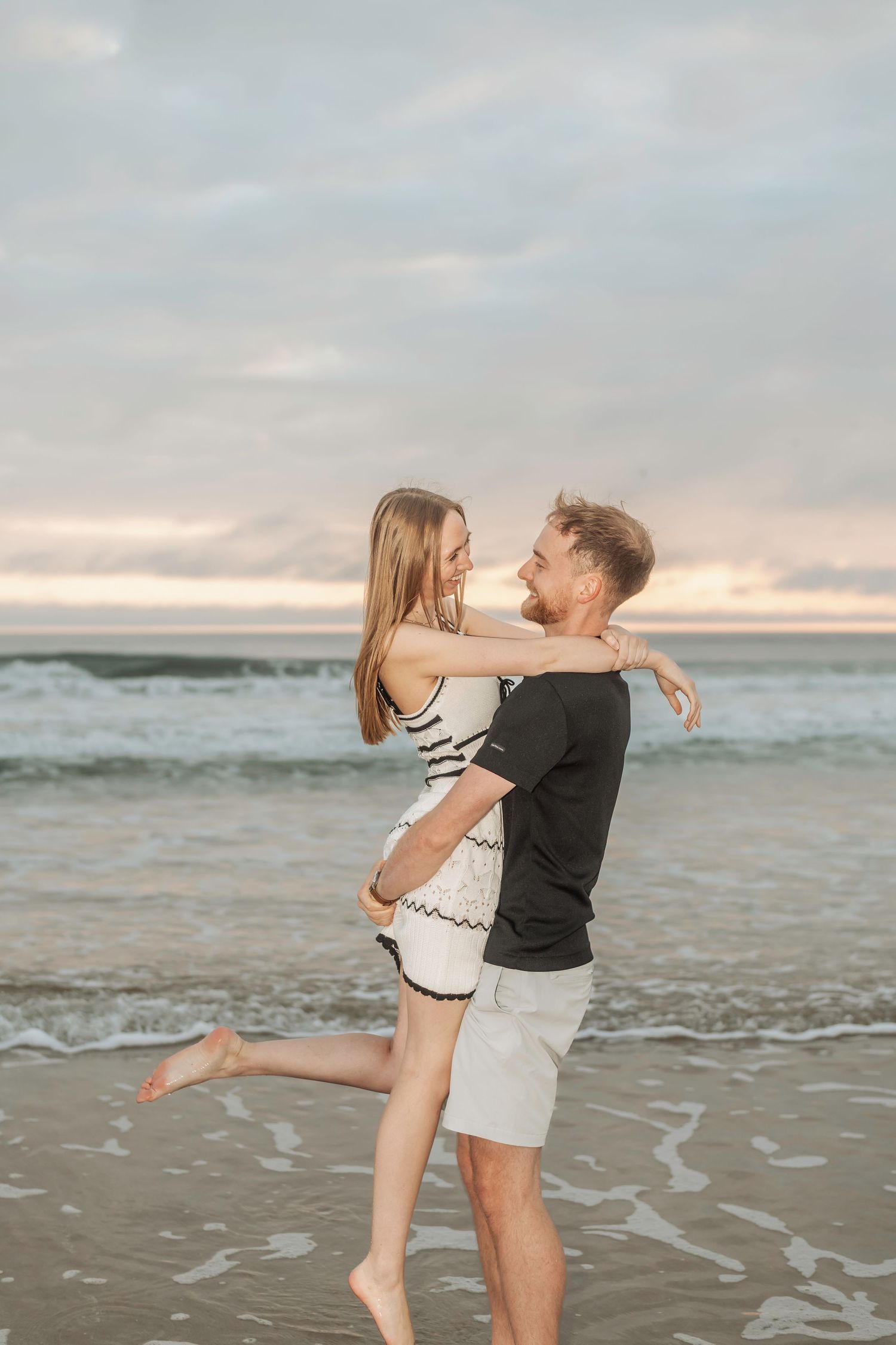 Couple embracing and smiling on a sandy beach at sunset, waves and pastel sky in the background.