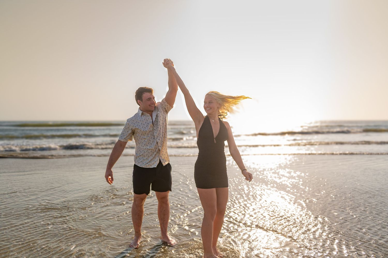 Couple dancing joyfully on a sunny beach at sunset, waves in the background.