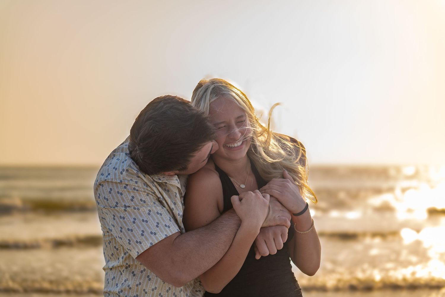 Couple embracing and laughing on a beach at golden hour sunset, woman smiling joyfully in romantic outdoor photo.