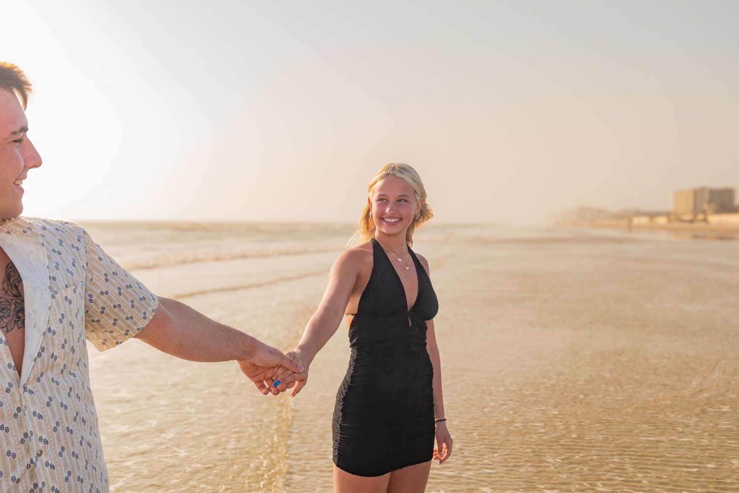 Couple holding hands on a sunny beach at golden hour, woman in black dress smiling joyfully.