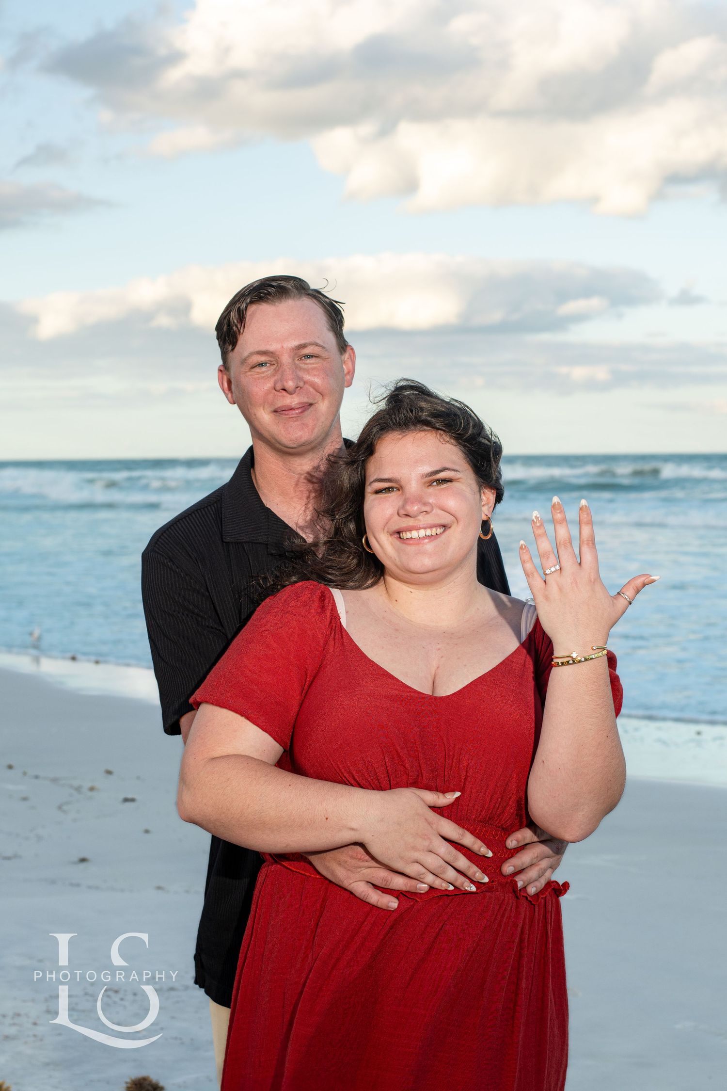 Happy couple posing on a beach at sunset showing off engagement ring.