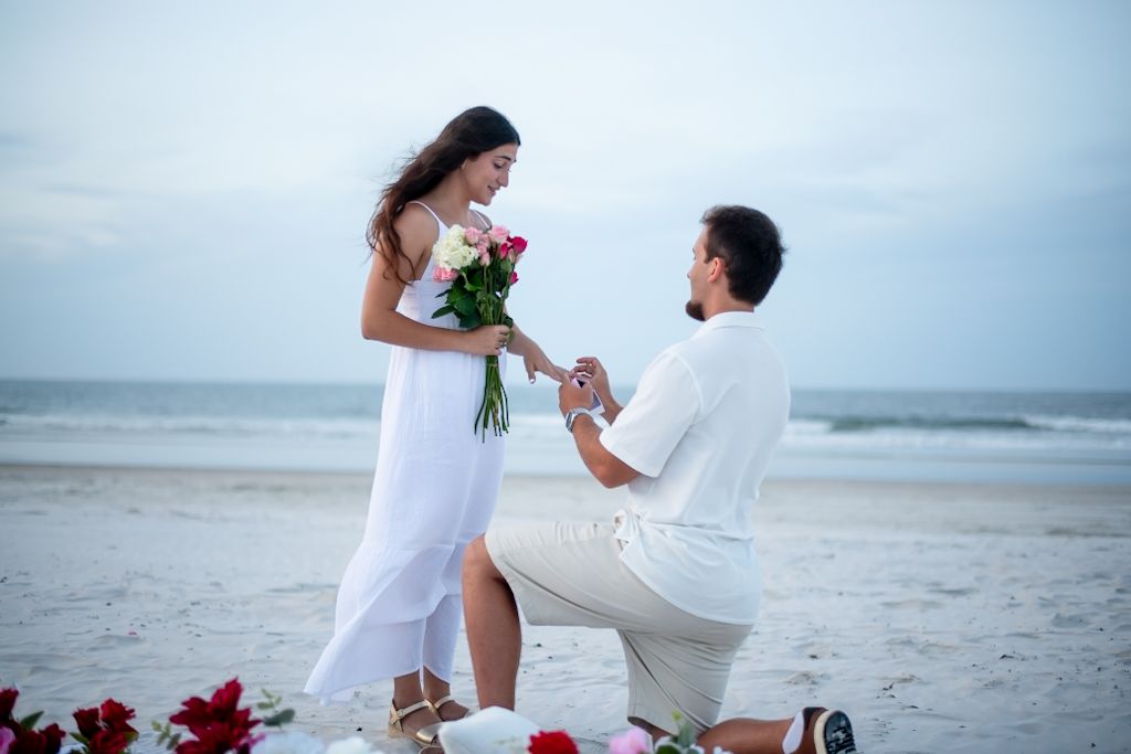Beach proposal scene with red roses scattered on white sand.