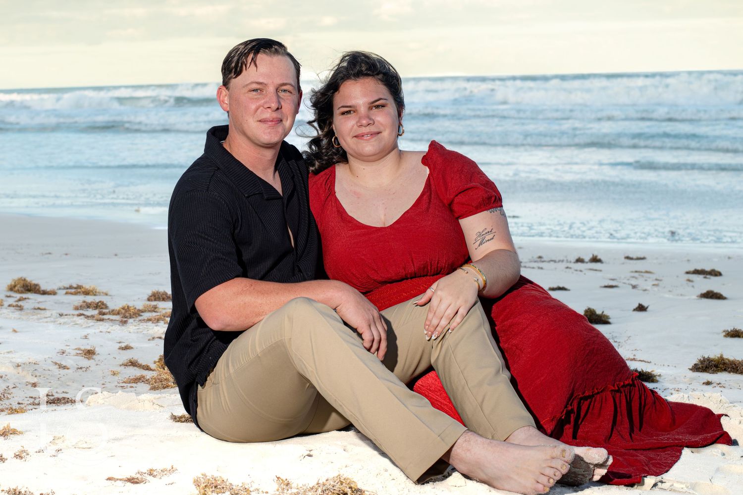 Couple in red and black attire sitting together on sandy beach.