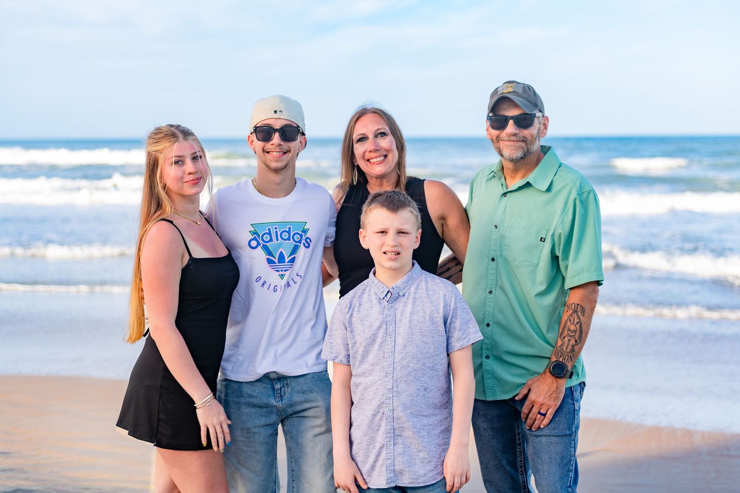 Family of five smiling together on a sunny beach with ocean waves in the background.
